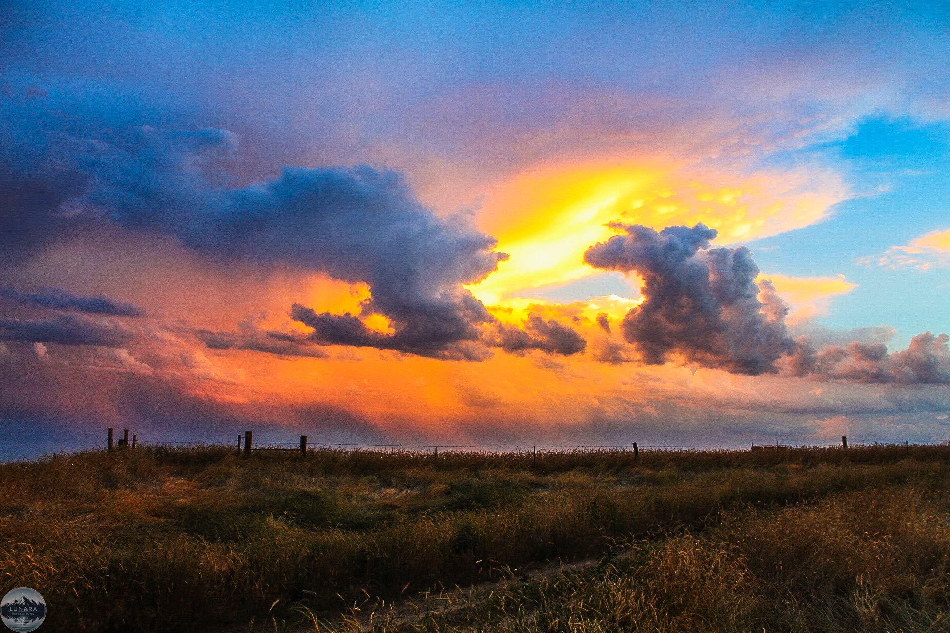 A scenic landscape of grassland with a fence in the foreground and a dramatic sunset sky with clouds in the background.