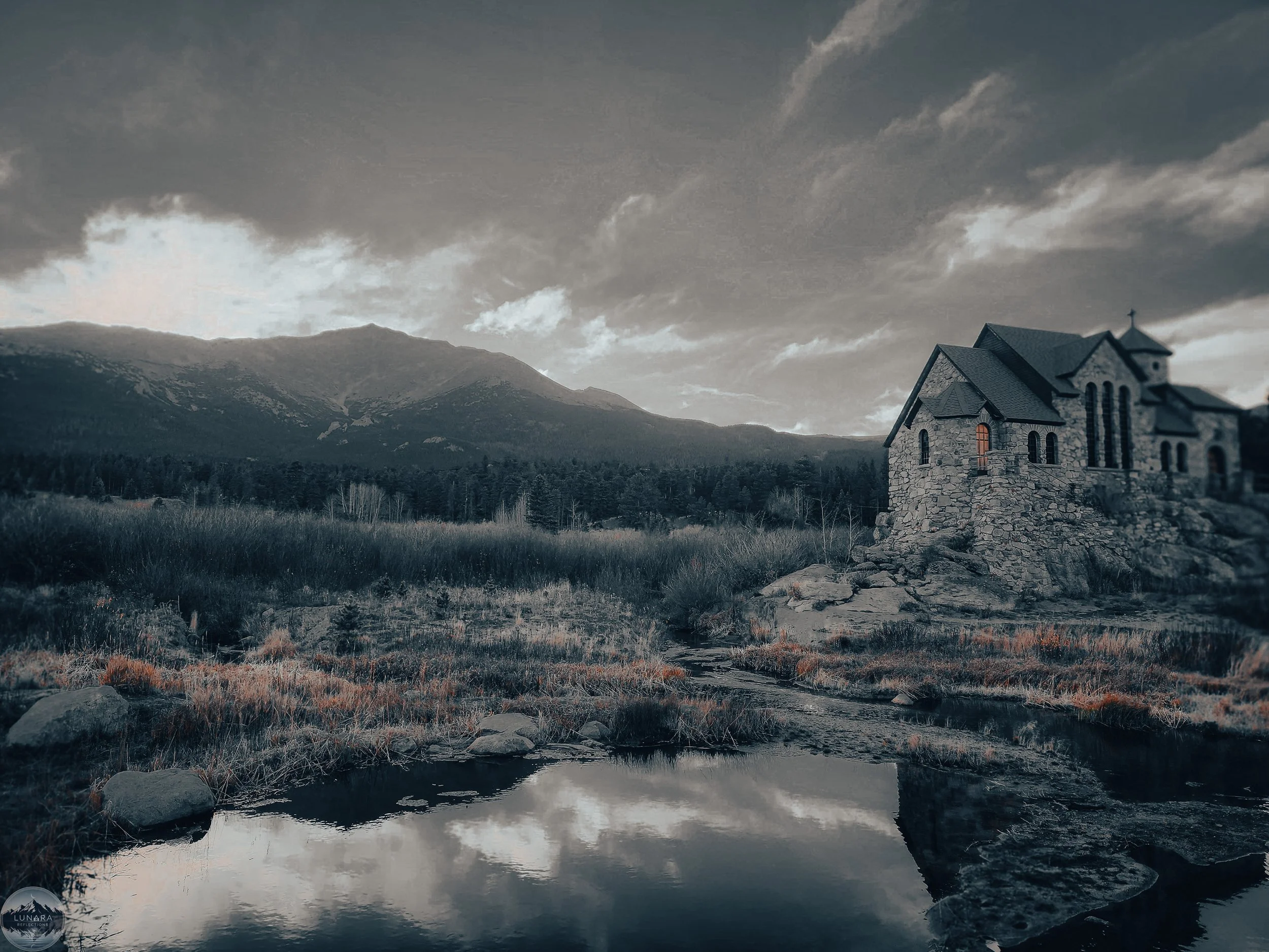 A black and white landscape featuring a hilltop church built with stone, overlooking a lake with rocks and grass, mountains, and a cloudy sky.