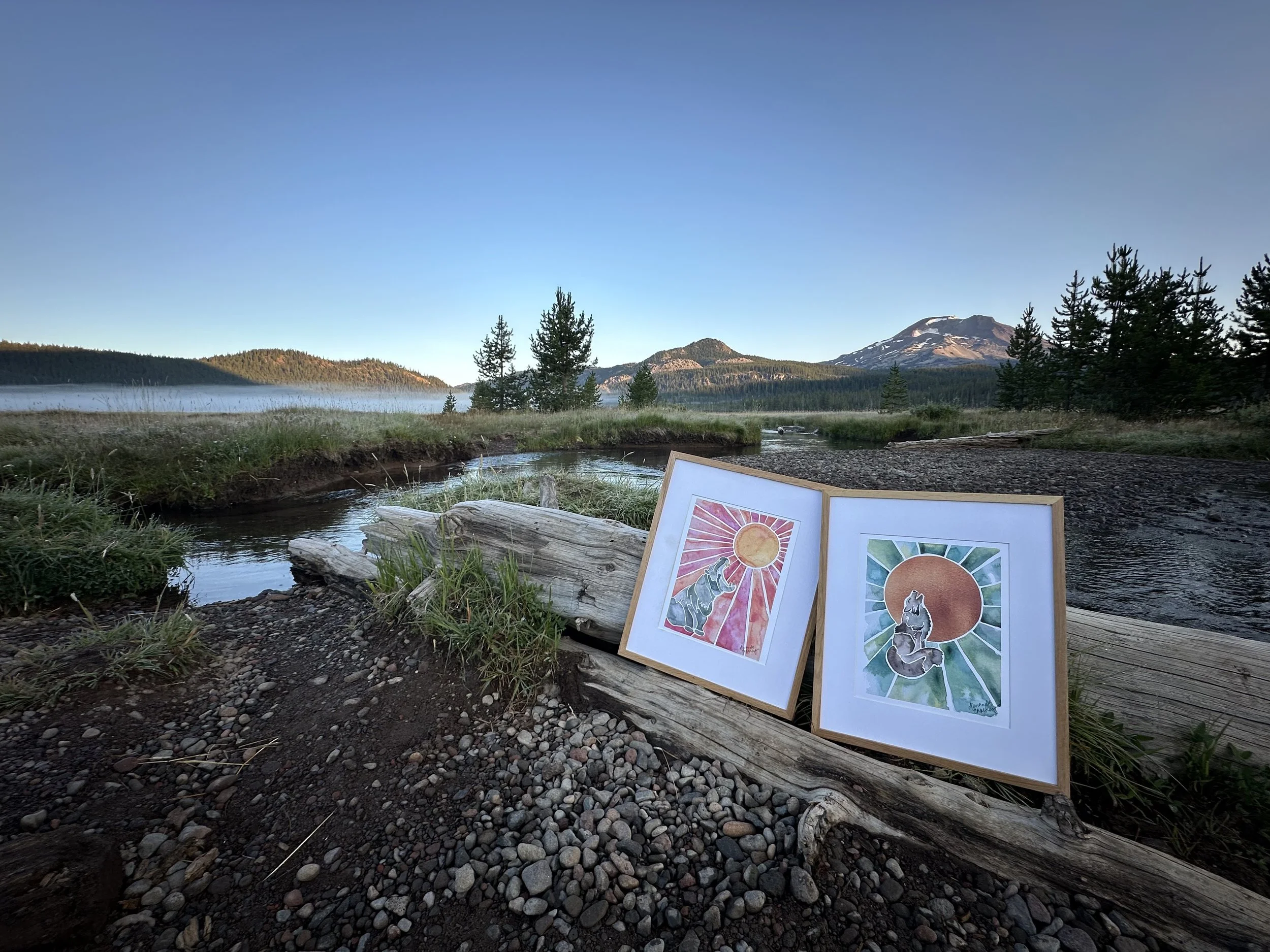 Two framed watercolor paintings of animals with a sun in the background, resting on a driftwood log by a river in a mountainous landscape during early morning or late evening.