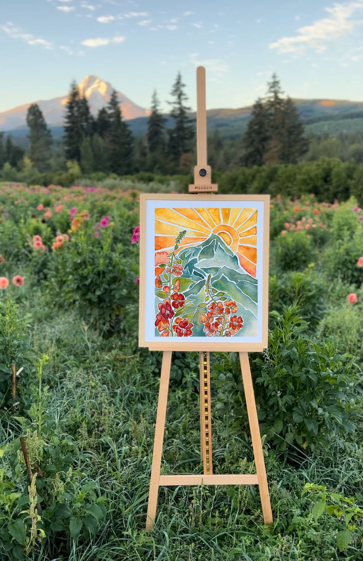 An easel displaying a colorful painting of a mountain, flowers, a sun, and rays of sunlight in a field with flowers, trees, and a mountain in the background under a partly cloudy sky.