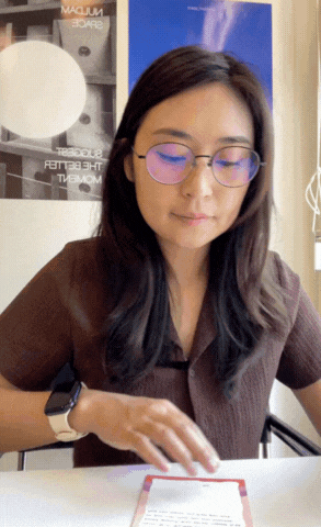 Woman with long dark hair and glasses reading a document at a table in an indoor setting.