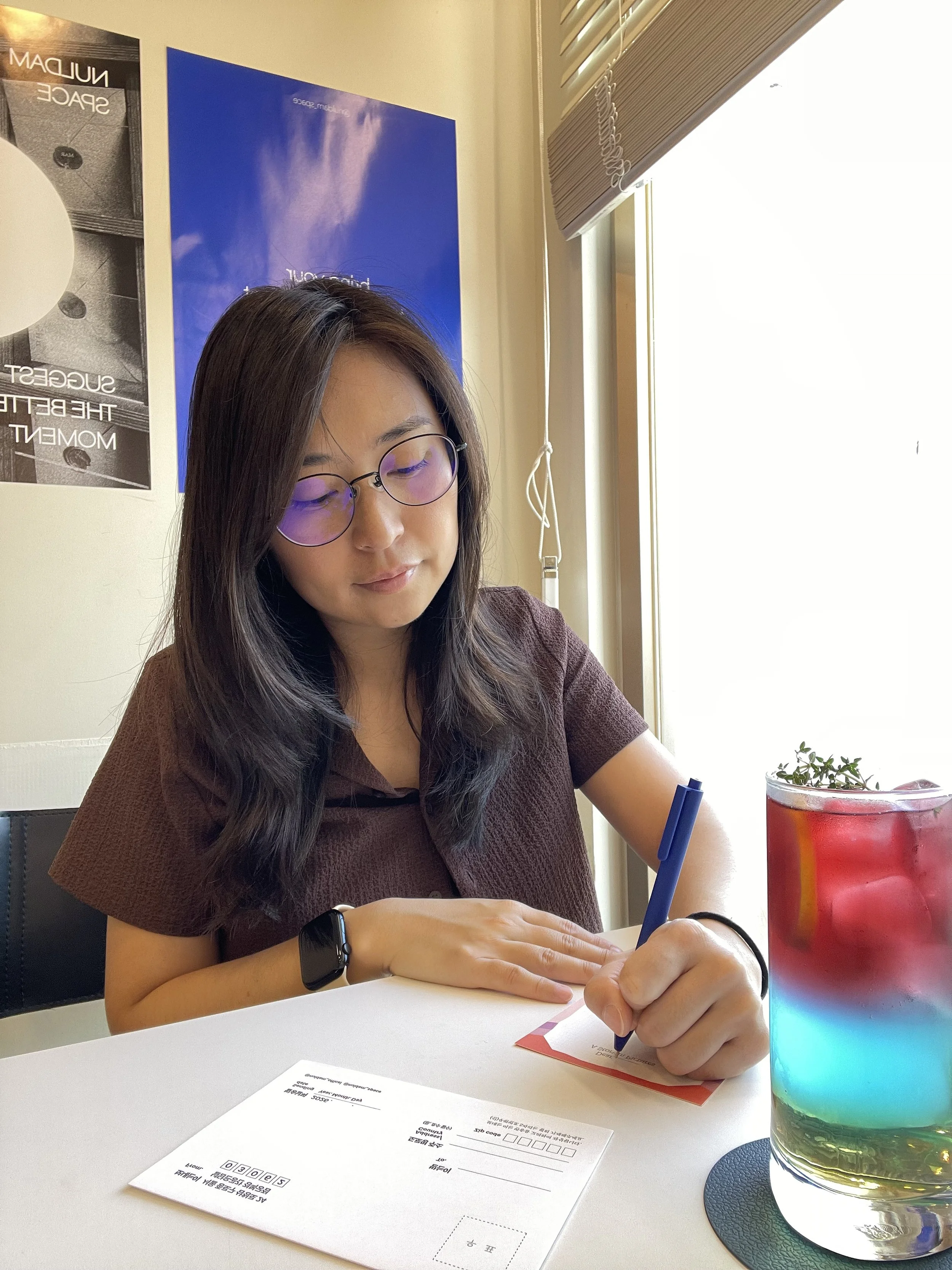 A woman with glasses writing on a piece of paper at a desk, with a colorful drink and posters on the wall behind her.