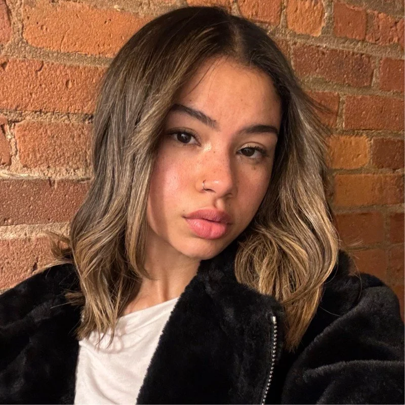 A young woman with wavy, shoulder-length hair, wearing a nose ring, a white top, and a black fuzzy jacket, standing in front of a brick wall.