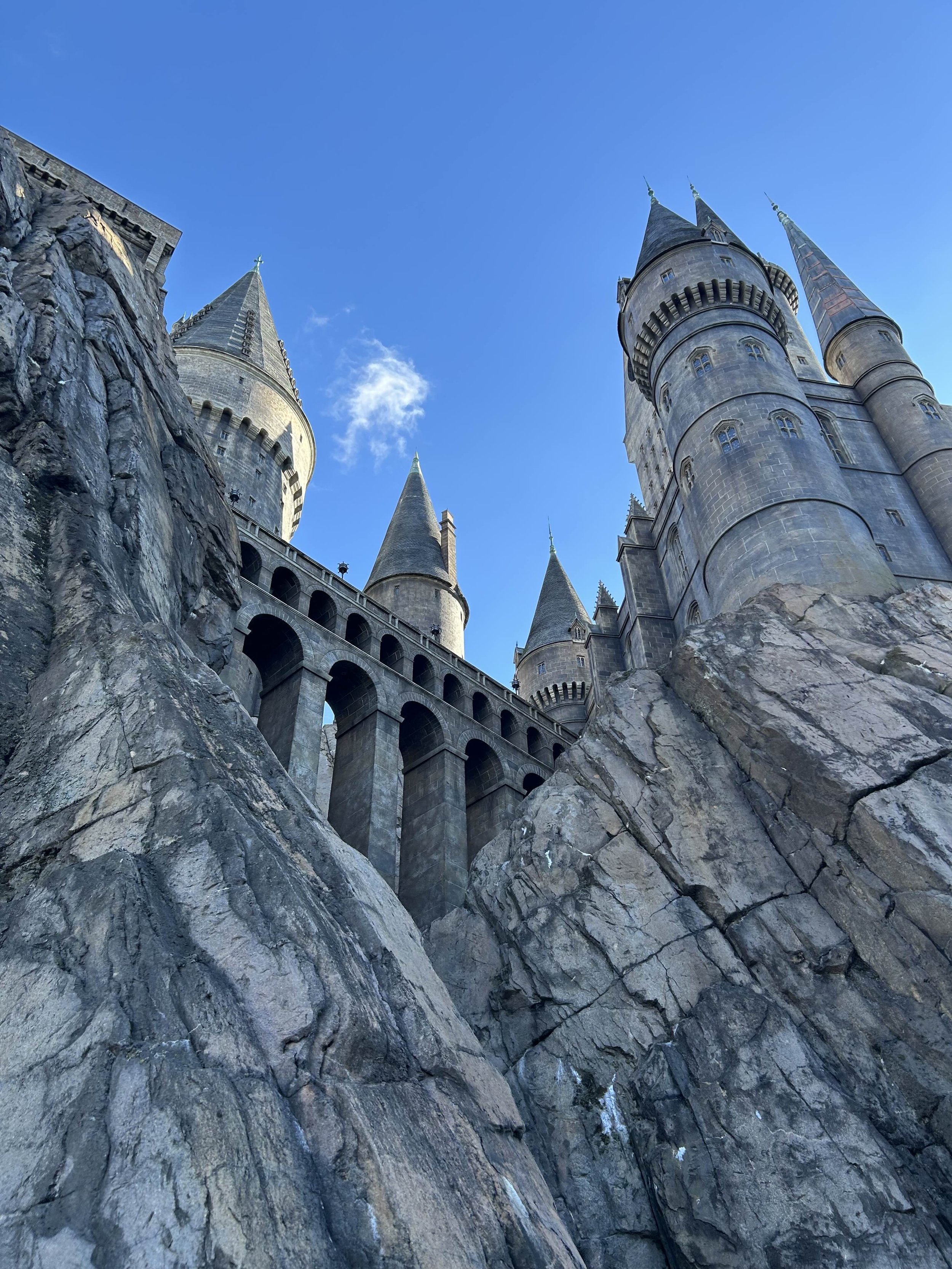 A low angle view of a castle with tall towers and pointed spires built on rocky terrain under a blue sky.