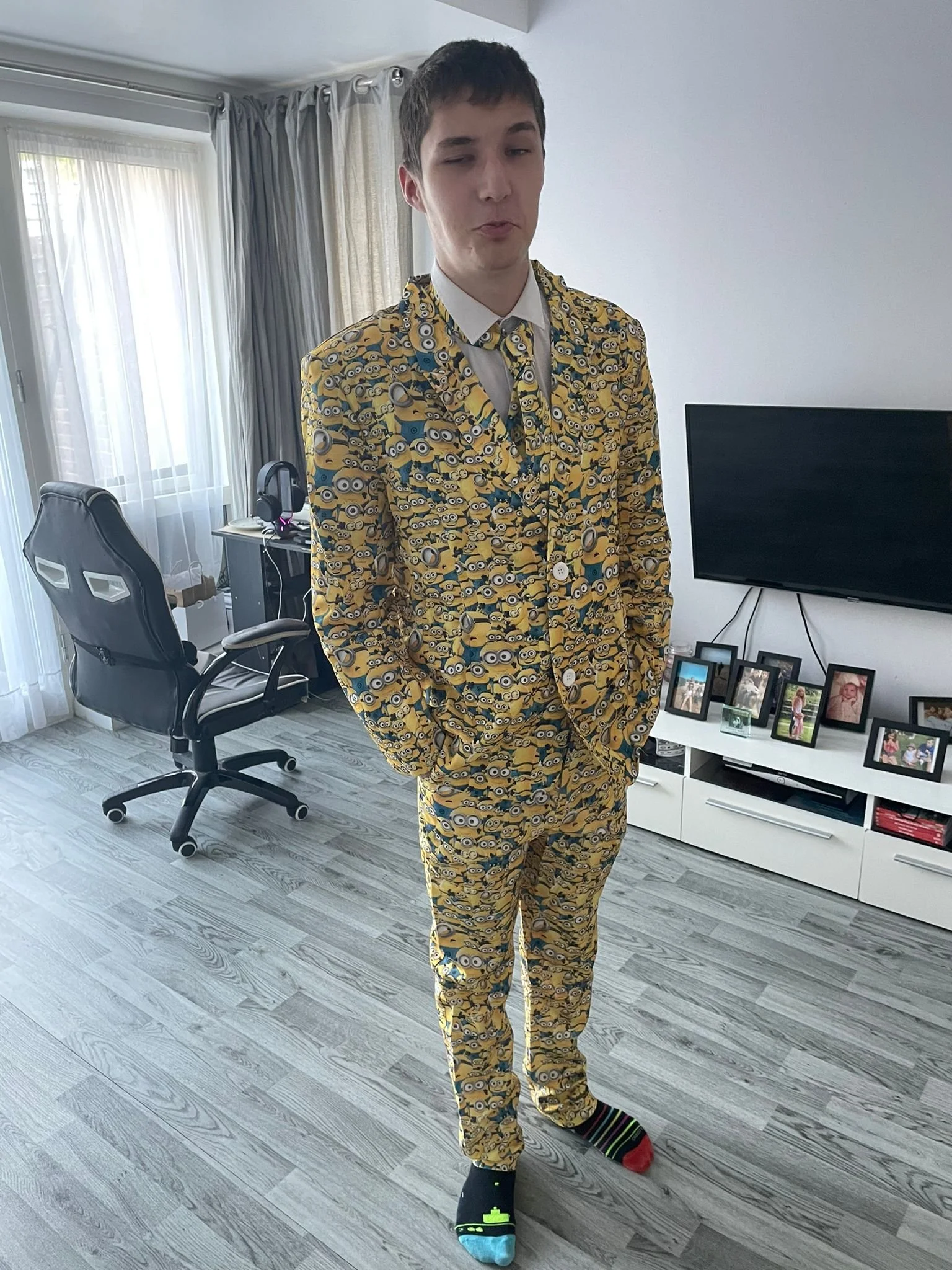 Young man in a Minions-themed suit with a white shirt, standing in a living room with a TV, picture frames, a desk, and an office chair.