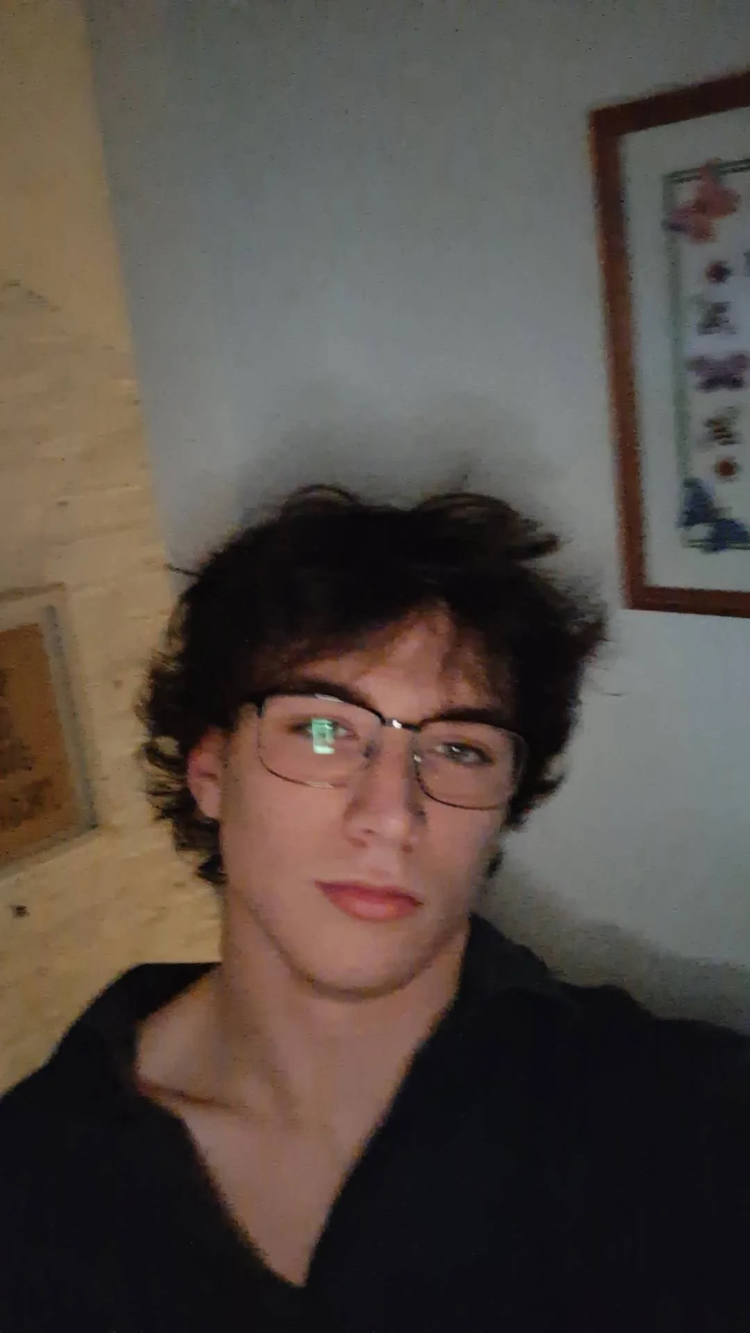 A young man with dark, wavy hair and glasses taking a selfie indoors, with a wall and framed artwork in the background.