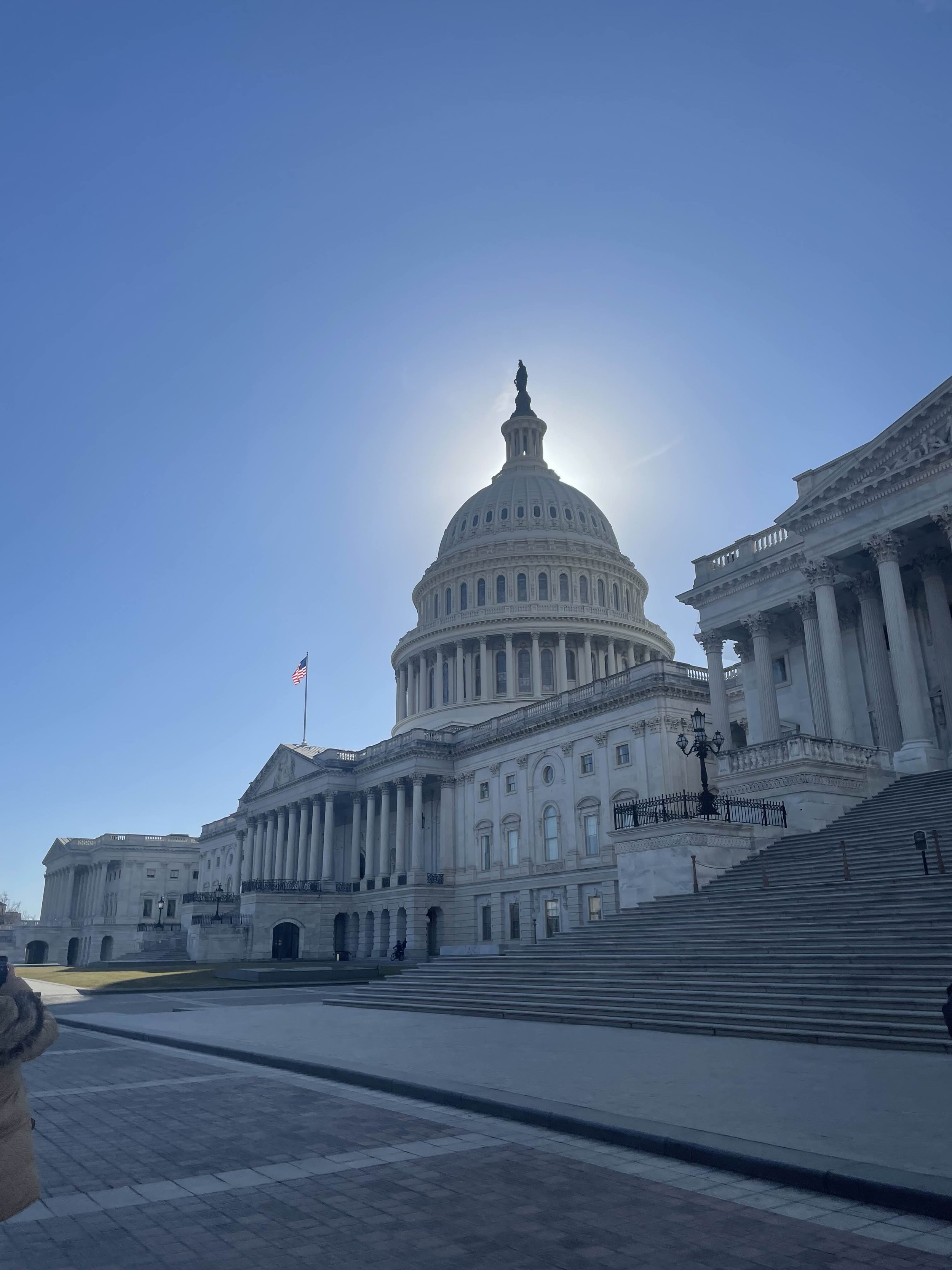 United States Capitol building on a clear day with the sun behind the dome, American flag flying, and stairs leading up to the entrance.