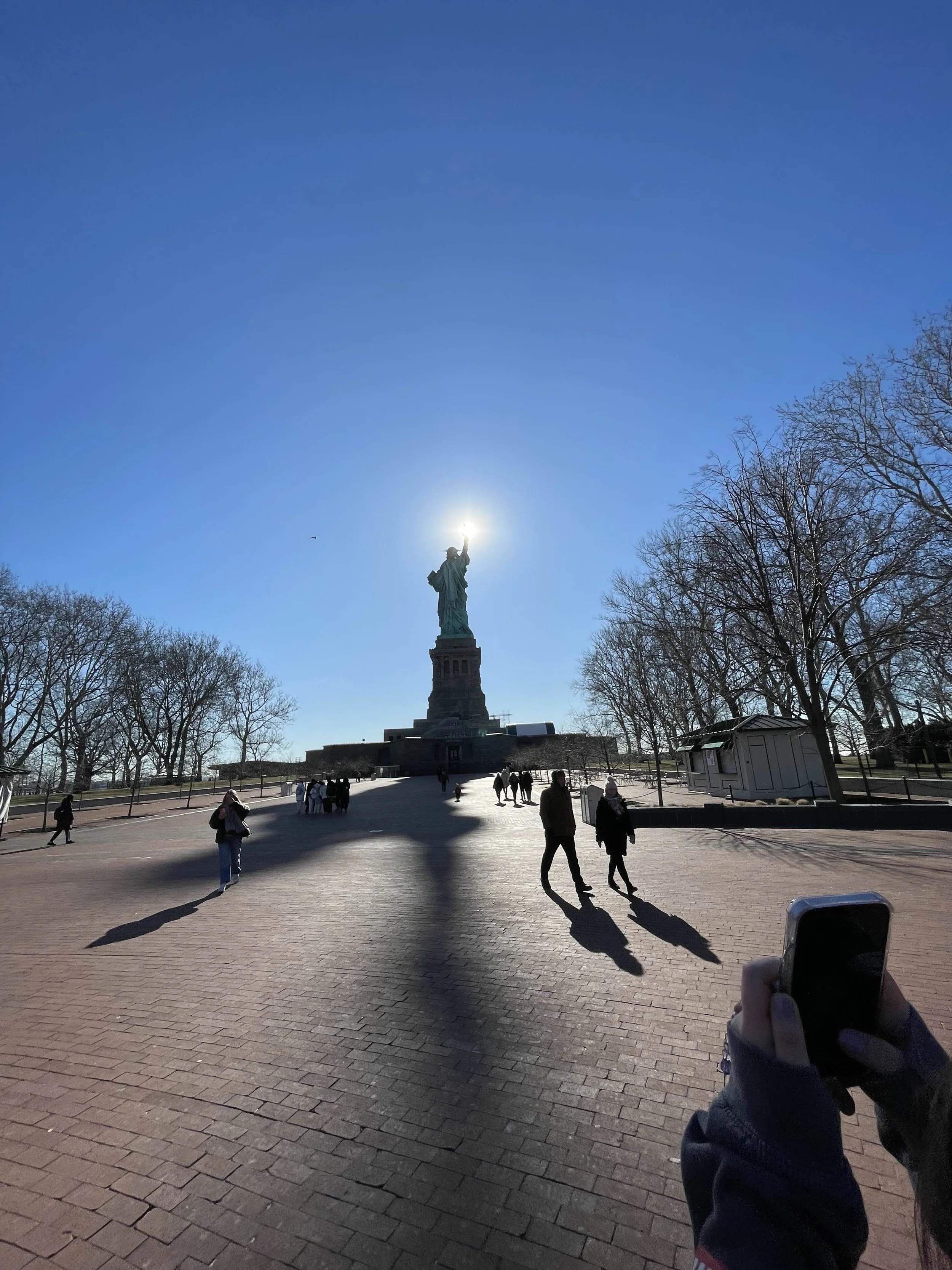 The Statue of Liberty in New York City with people walking around on a paved area, trees on the sides, and the sun shining behind the statue's torch.