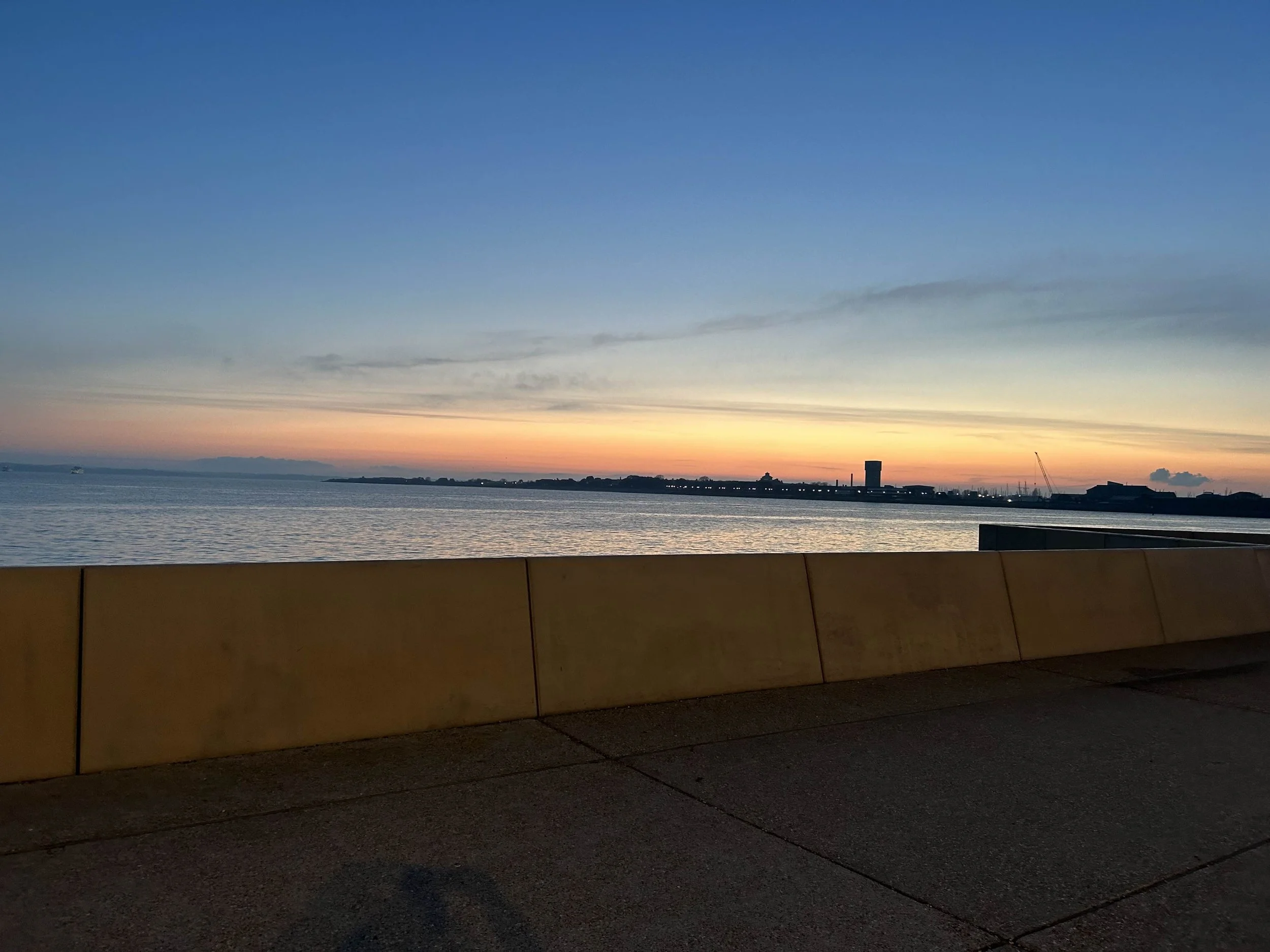 Portsmouth coast, Sunset over a body of water with a distant city skyline and a protected walkway in the foreground with the sun setting.