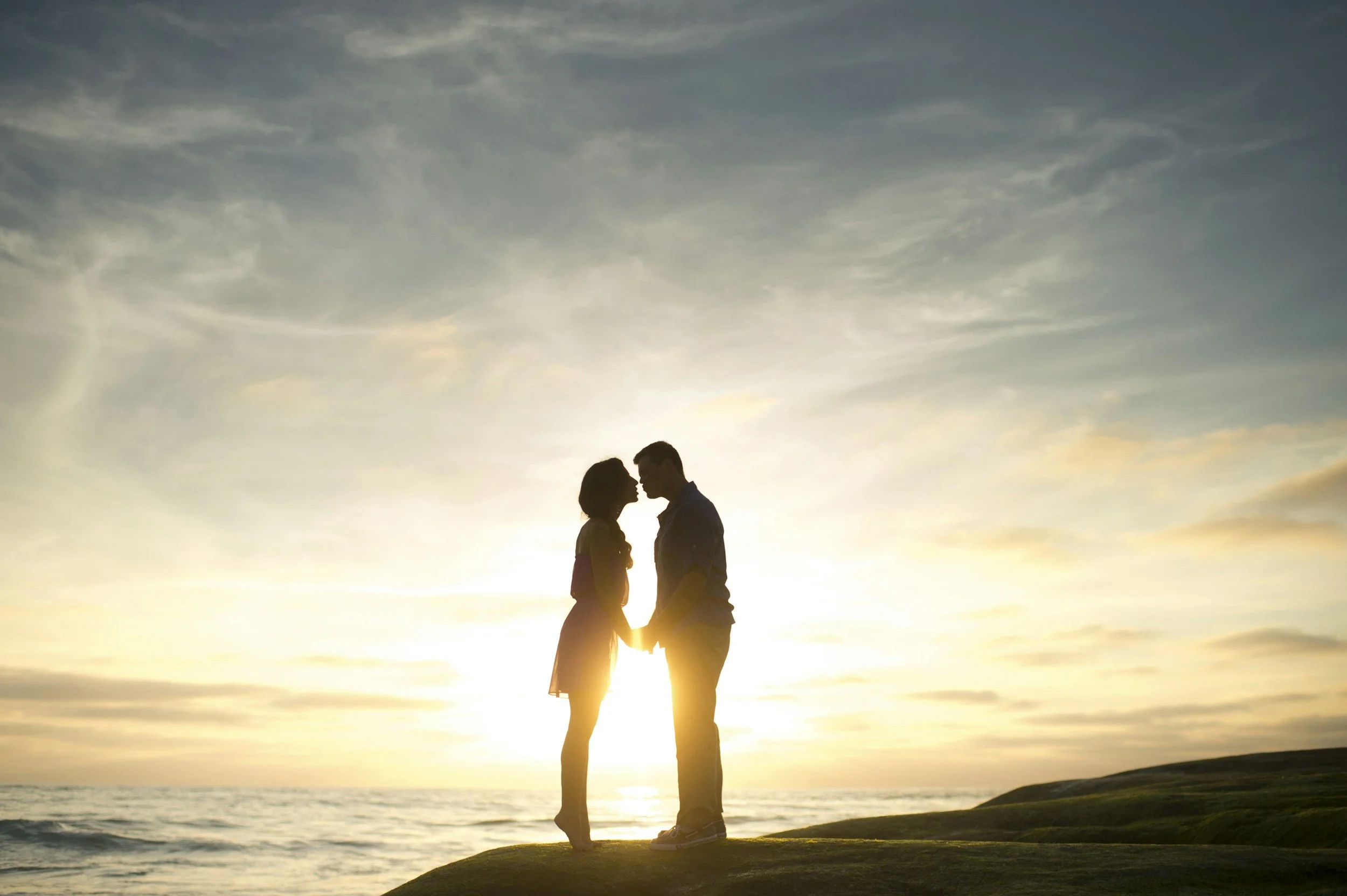 Silhouette of a couple holding hands and touching foreheads on a grassy shore during sunset.