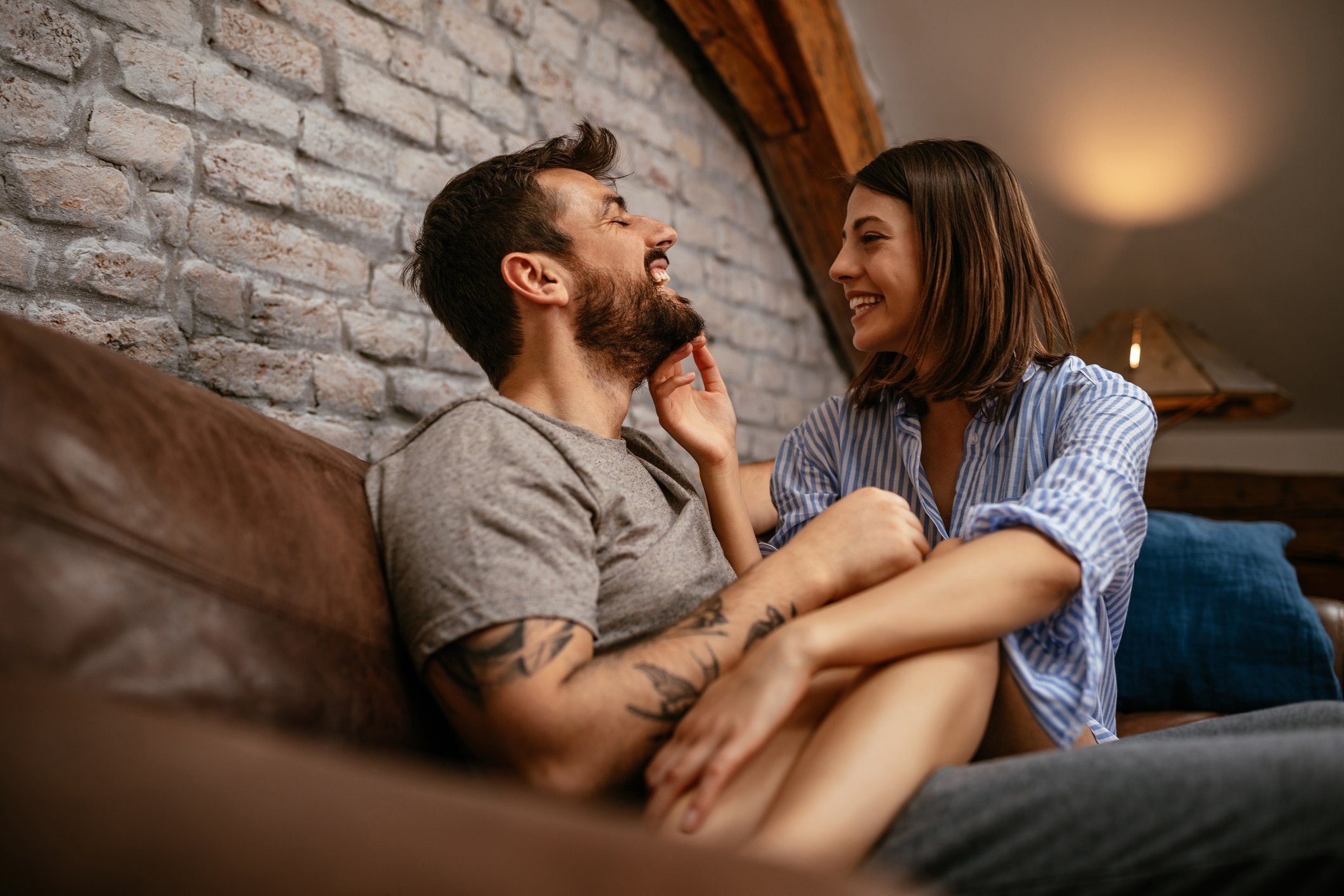 A happy couple sitting on a couch, smiling and enjoying each other's company, in a cozy room with a brick wall background.