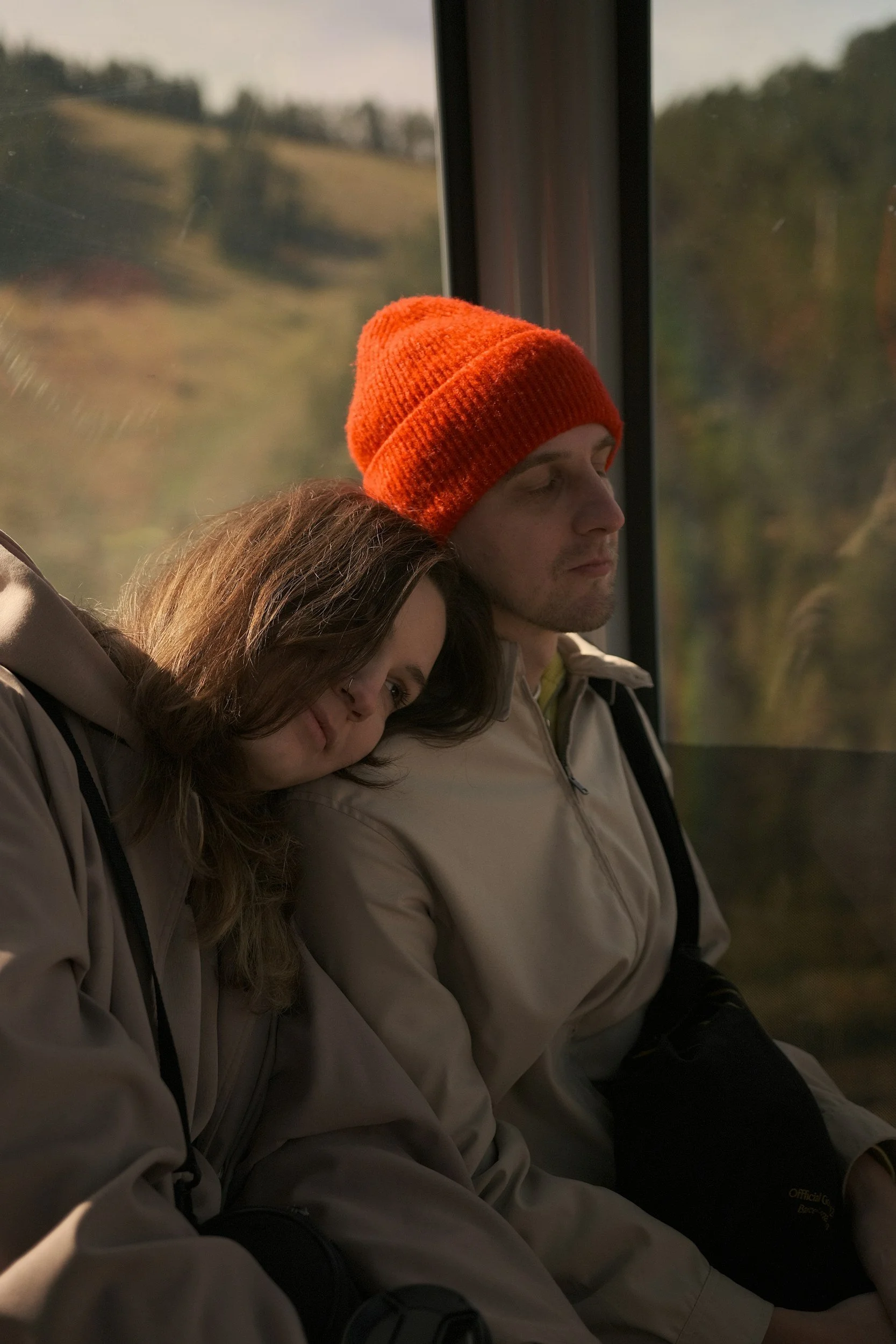 A young woman resting her head on a young man's shoulder on a bus or train, with a scenic landscape visible through the window behind them.