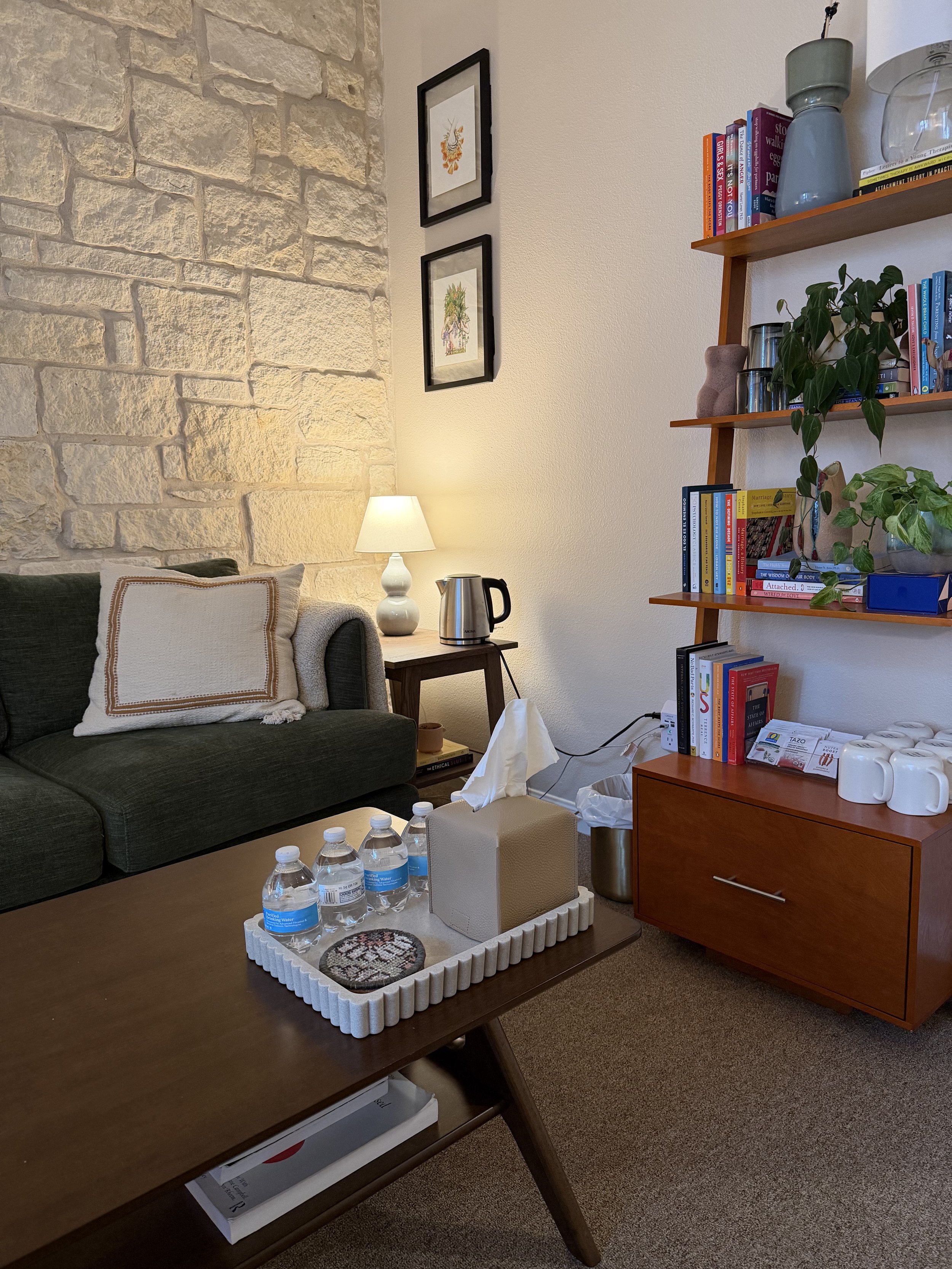 Apego Therapy's office resembles a living room with stone accent wall, green sofa, white pillow, wooden side table with lamp, water bottles on tray, tissue box, books and plants on wooden shelves, and framed artwork on wall.