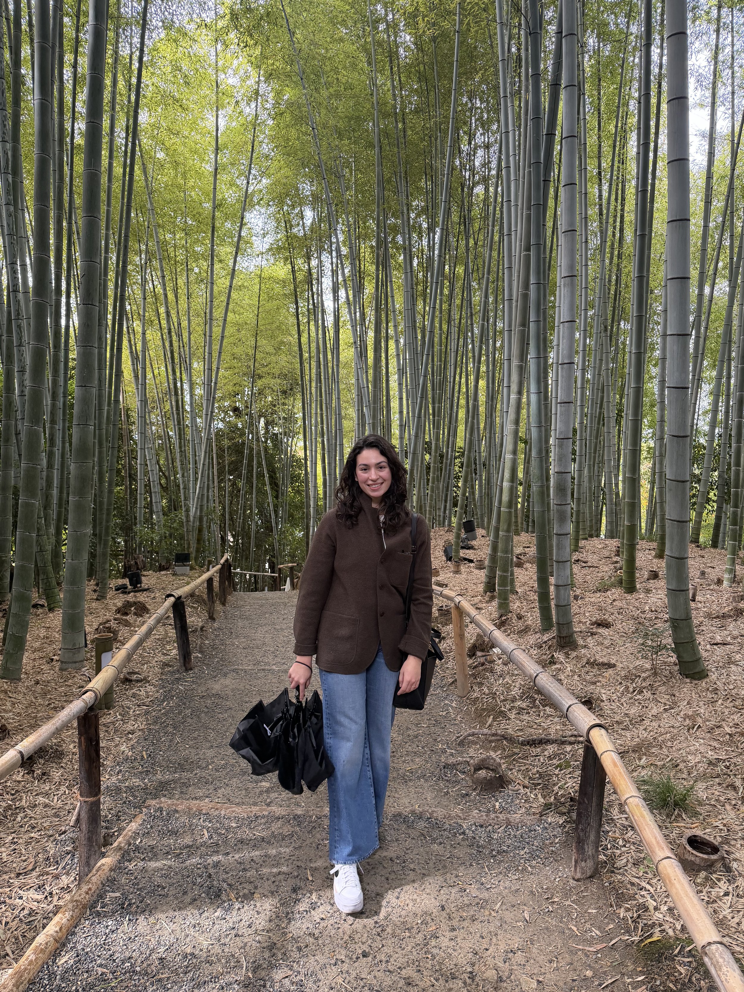 Maria Mohamed walking on a dirt path in a bamboo forest, holding black shopping bags, smiling, wearing a brown jacket, light blue jeans, and white sneakers.