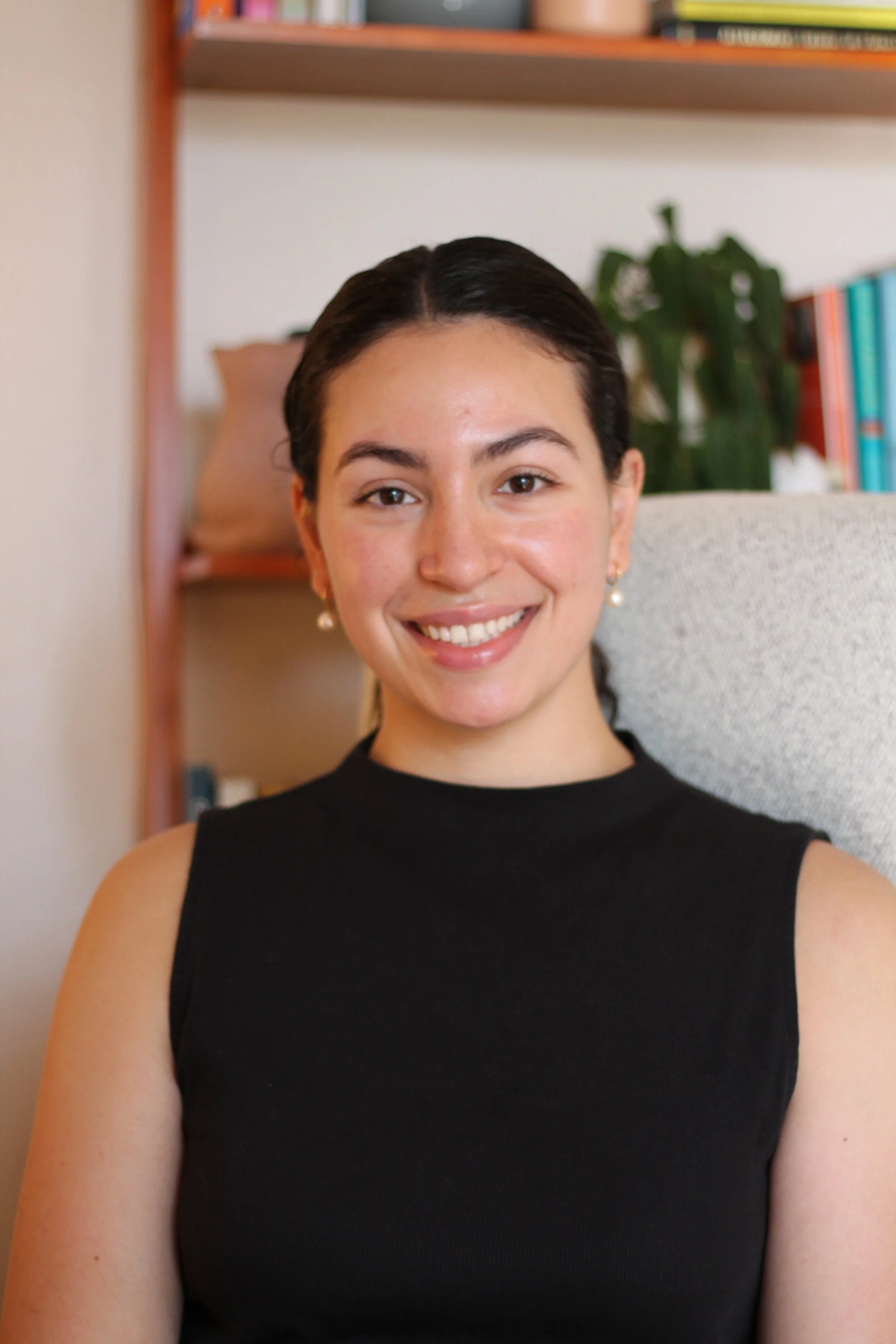 Maria Mohamed, a couples and individual therapist offering services in Austin. She is pictured with dark hair and earrings smiling, sitting in a room with books and a plant in the background.