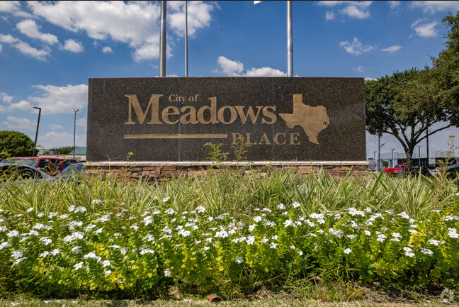 City of Meadows Place sign with trees and cars in the background under a partly cloudy sky.