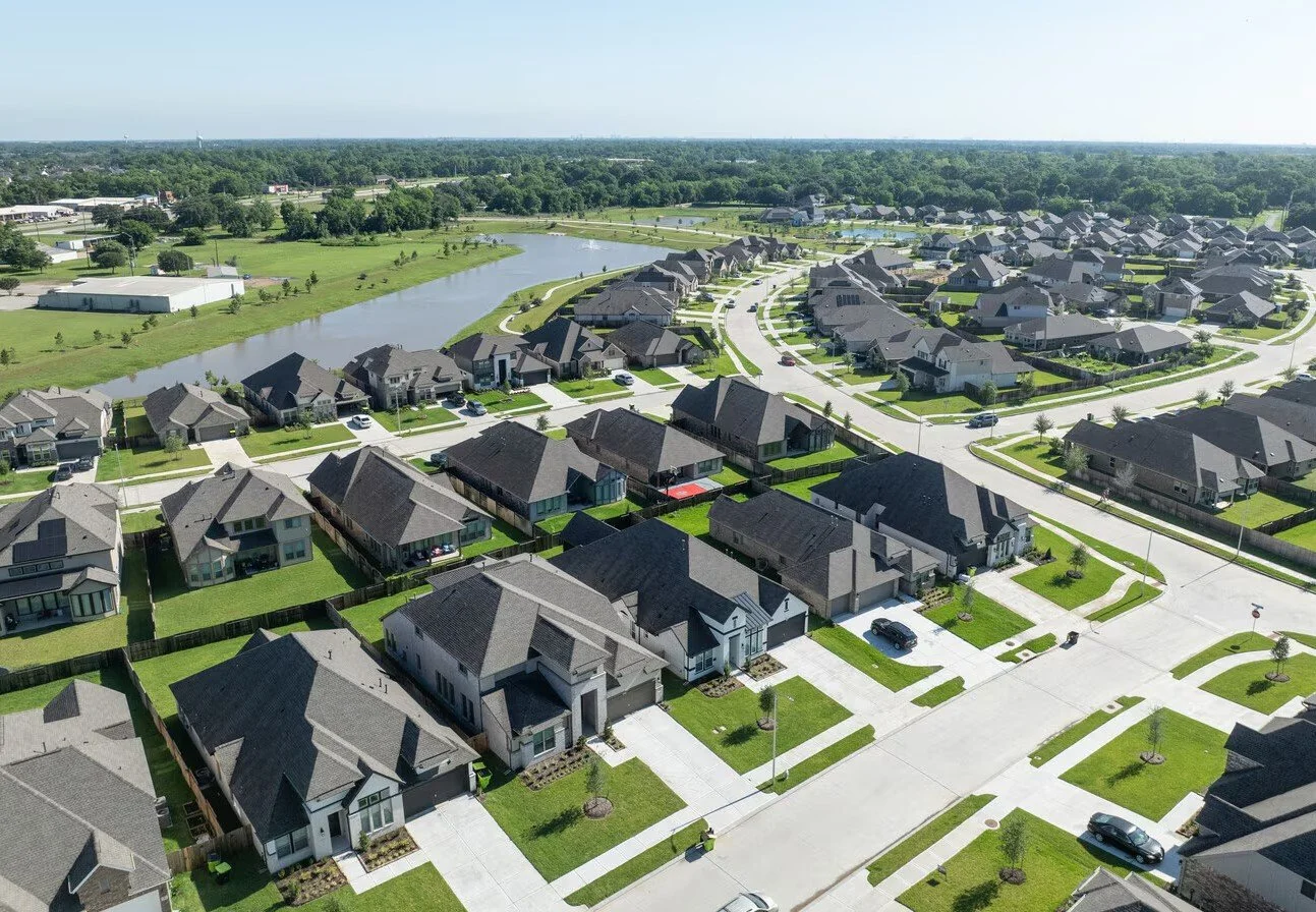 Aerial view of a suburban neighborhood with single-family houses, well-kept lawns, and streets. A pond is visible in the background with trees surrounding it.