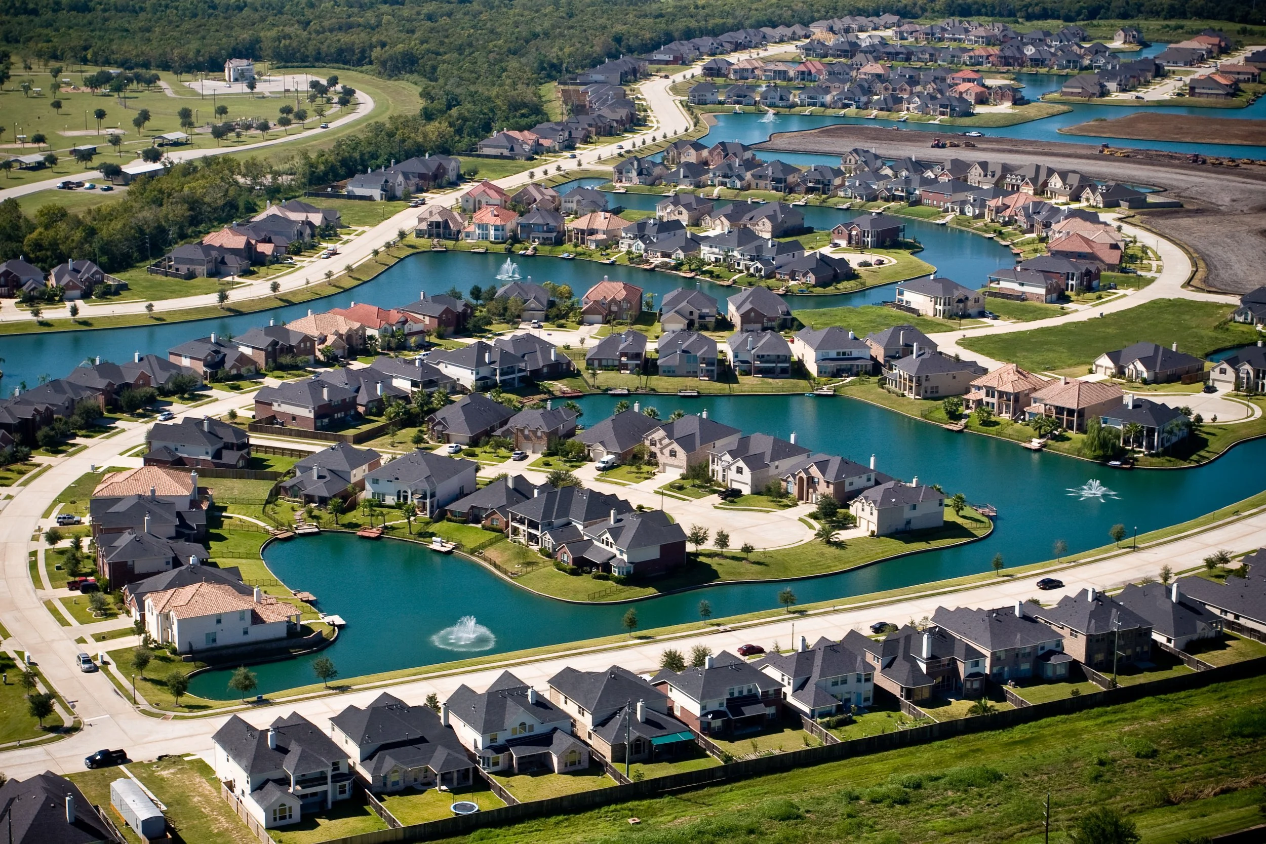 Aerial view of a suburban residential neighborhood with lakes, houses, and greenery, featuring multiple water fountains and winding roads.