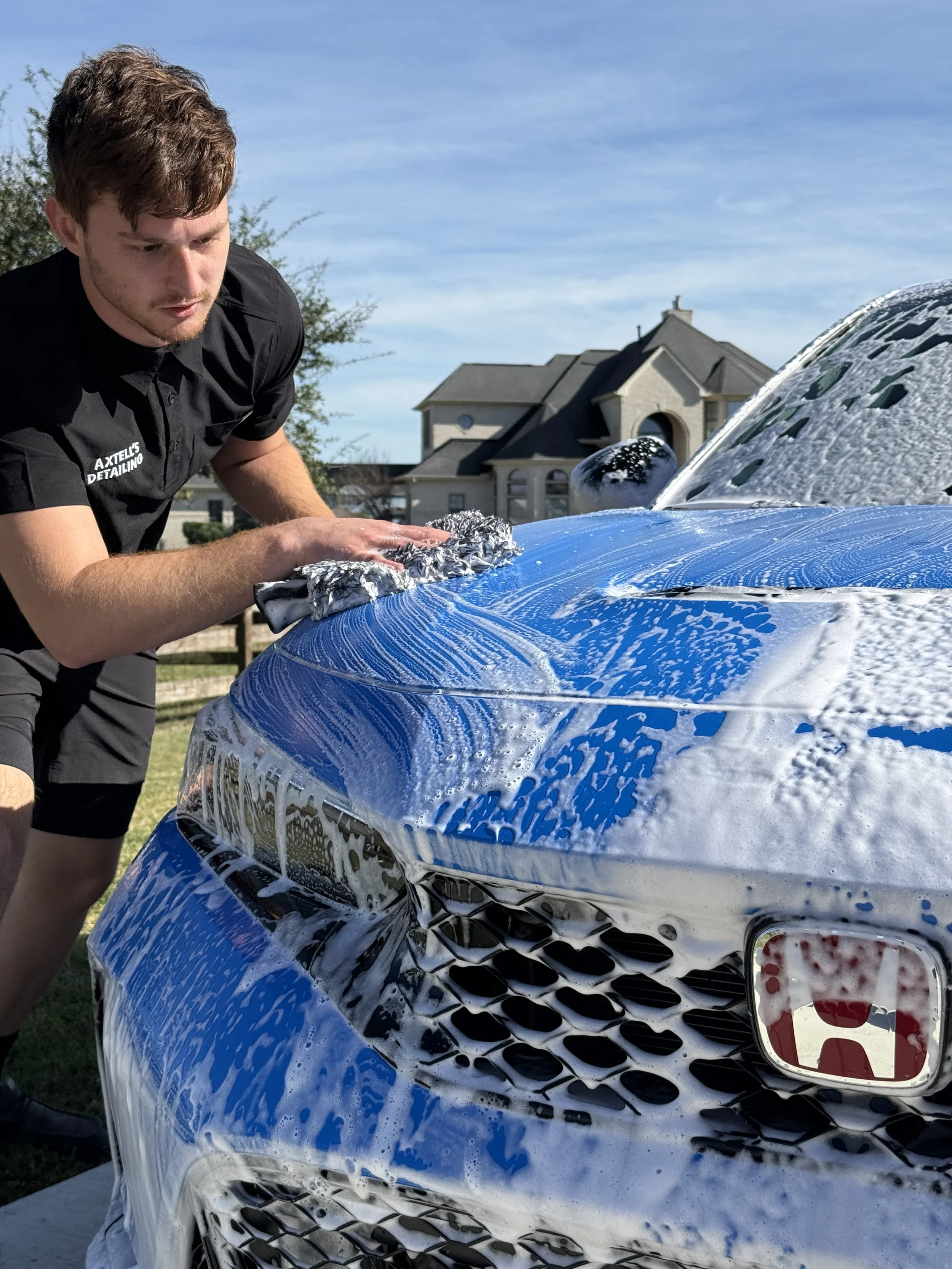 A man washing a blue car with soap and water, scrubbing the hood with a cloth, outdoors on a sunny day with house in background.