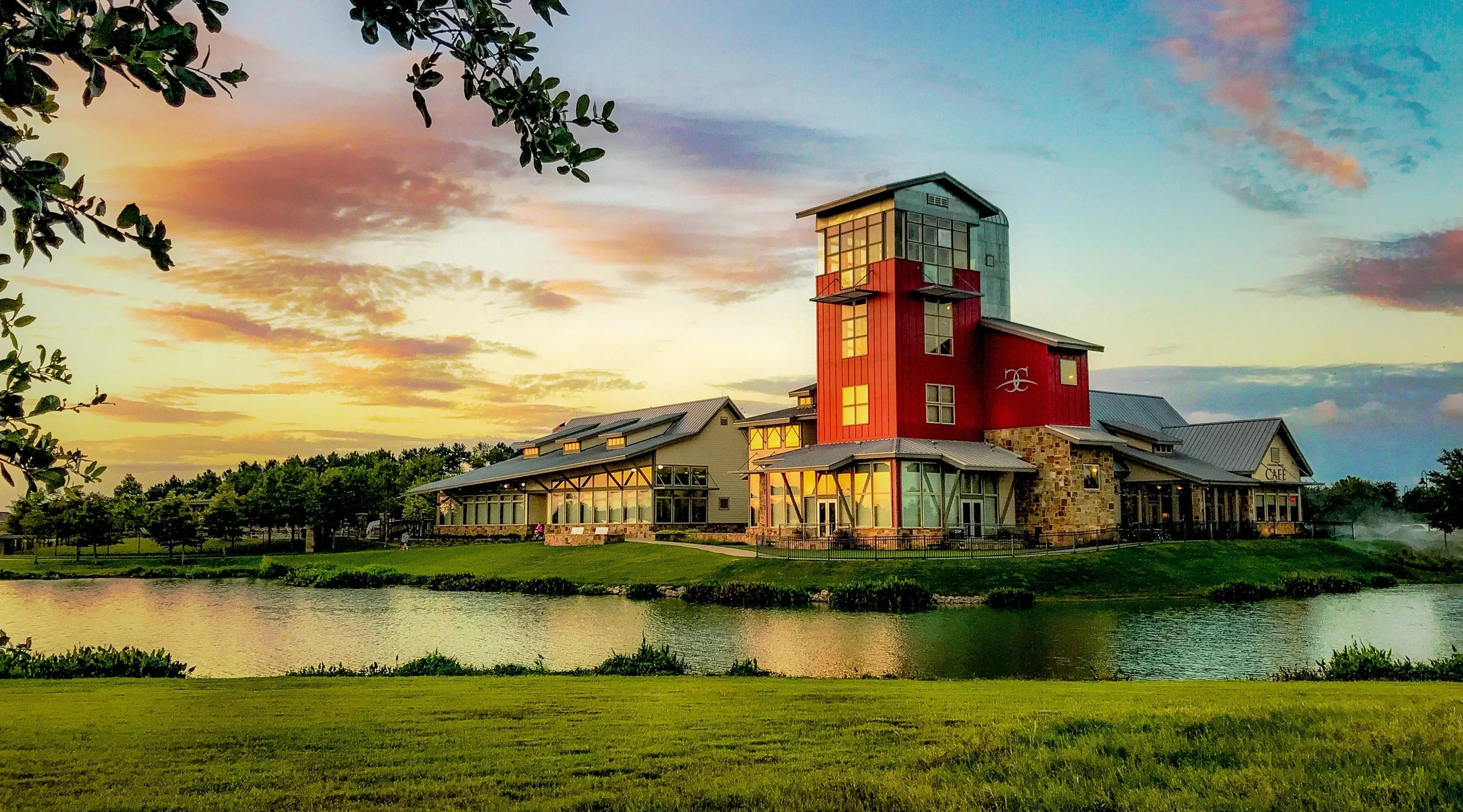 A large modern building with a red tower, glass windows, and stone accents, set beside a body of water during sunset with a colorful sky.