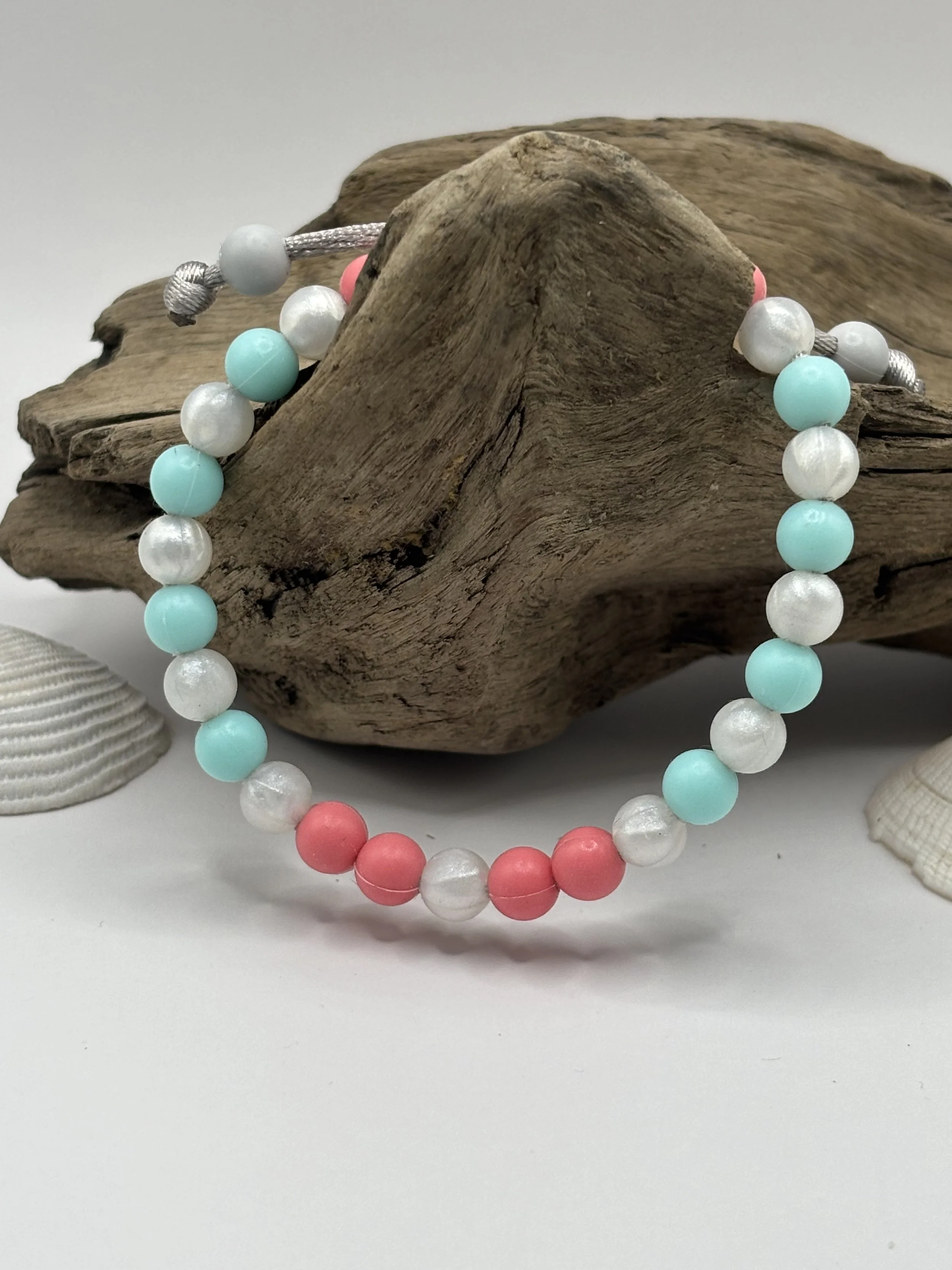 Colorful beaded bracelet with pink, white, and turquoise beads draped over a driftwood piece, with seashells in the background on a white surface.