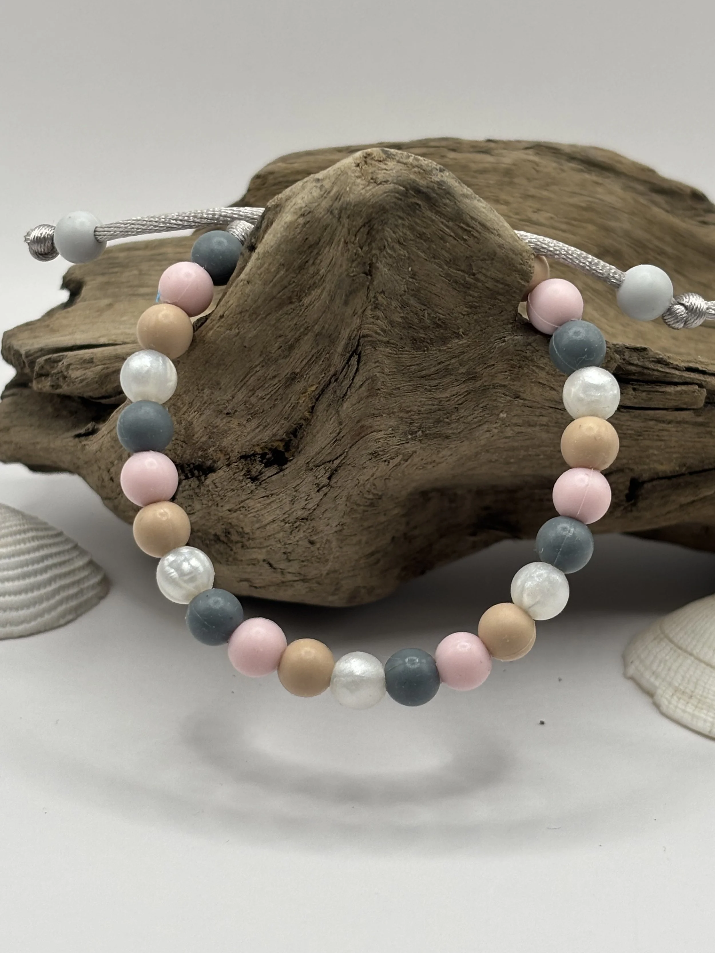 Beaded bracelet with pastel pink, cream, white, blue, and gray beads on a silver adjustable cord, draped over a piece of driftwood with seashells nearby.