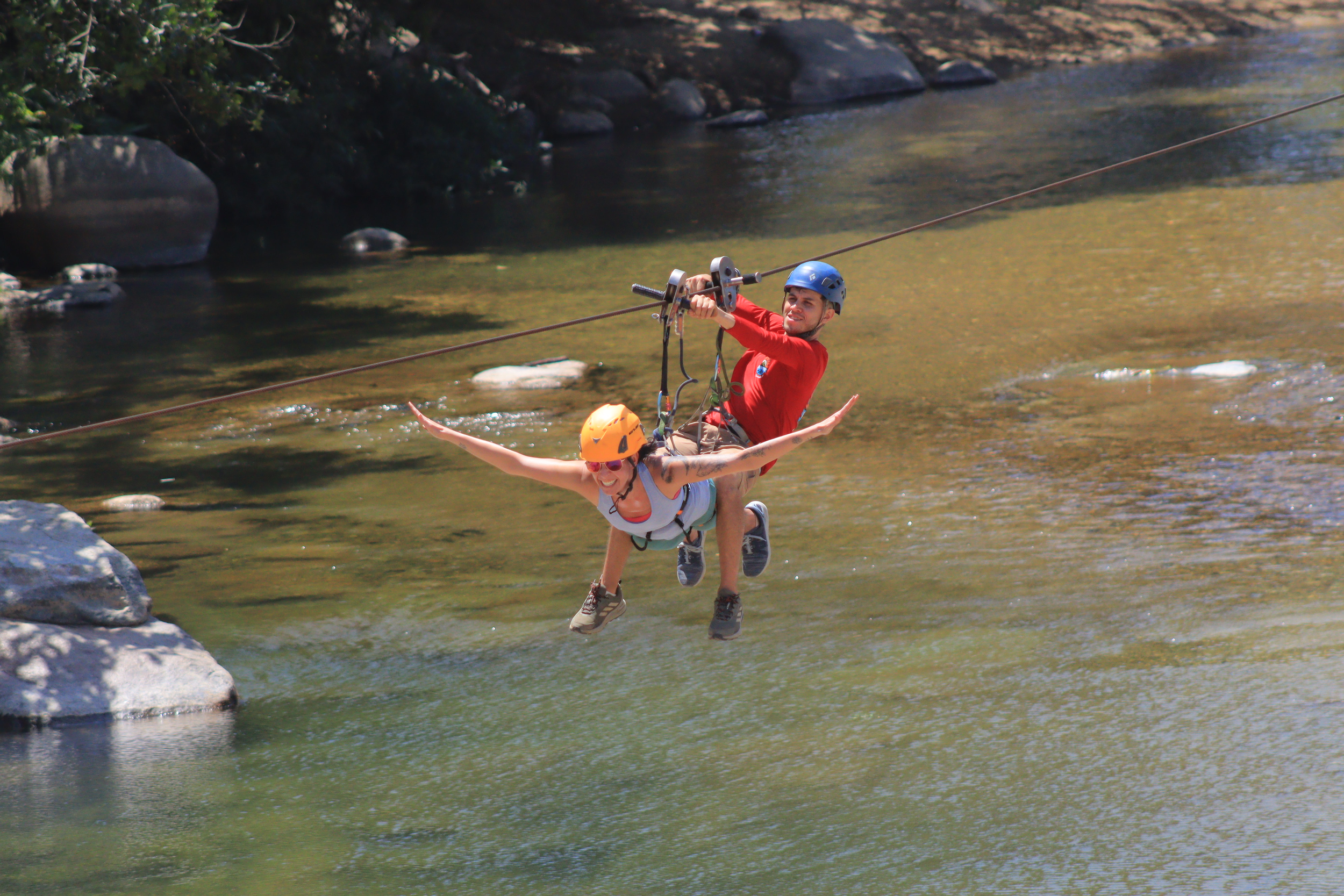 Two people, a man and a woman, ziplining over a river; the woman is in front with arms outstretched and a big smile, wearing an orange helmet, sunglasses, and a tank top, while the man is behind her holding the zipline harness, wearing a blue helmet and red shirt.