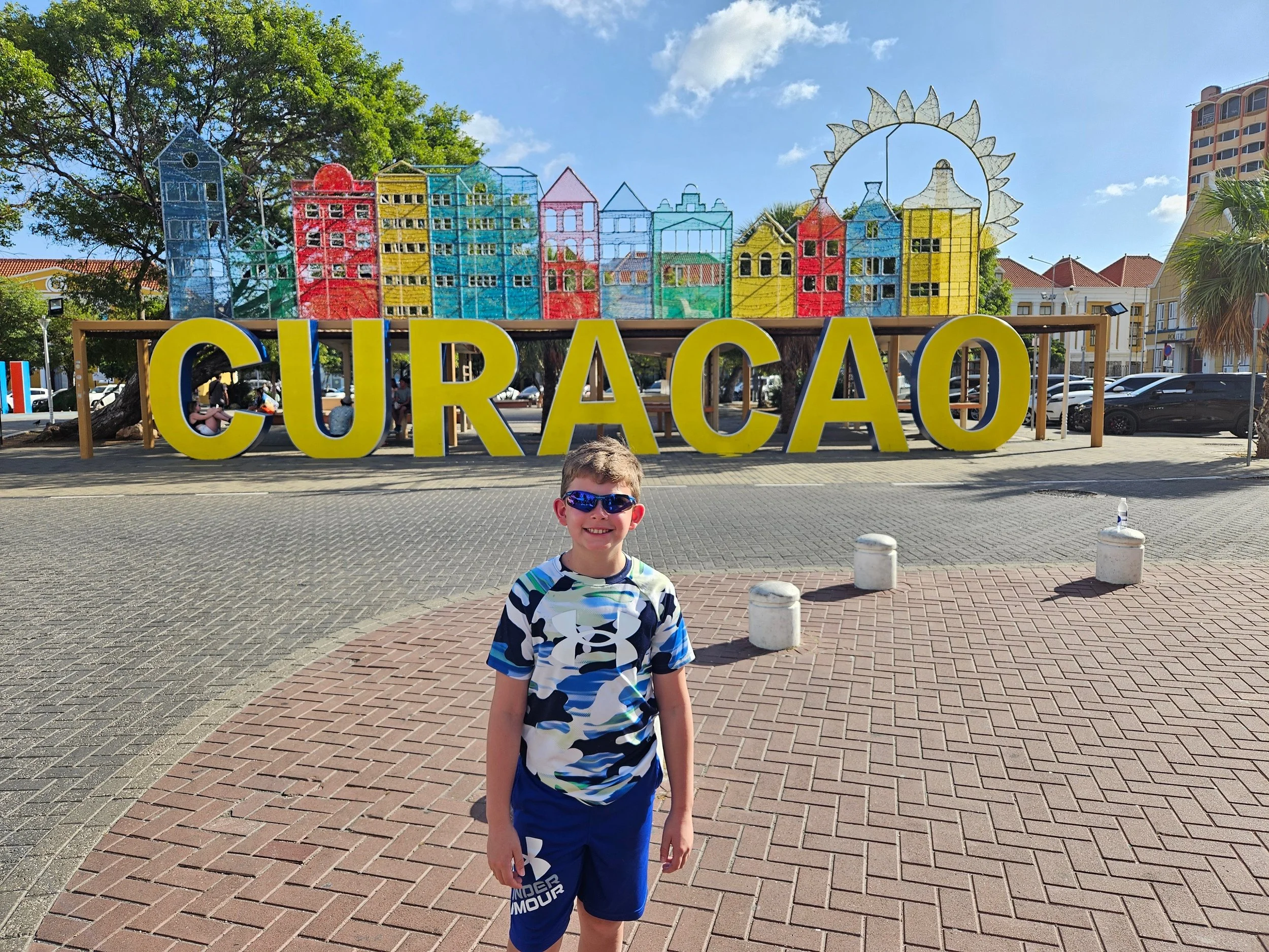A smiling boy wearing sunglasses, a camouflage shirt, and blue shorts stands in front of a large yellow 'CURAÇAO' sign. Behind the sign, there are colorful buildings and a decorative sun and buildings sculpture, with a tree, parked cars, and buildings in the background.