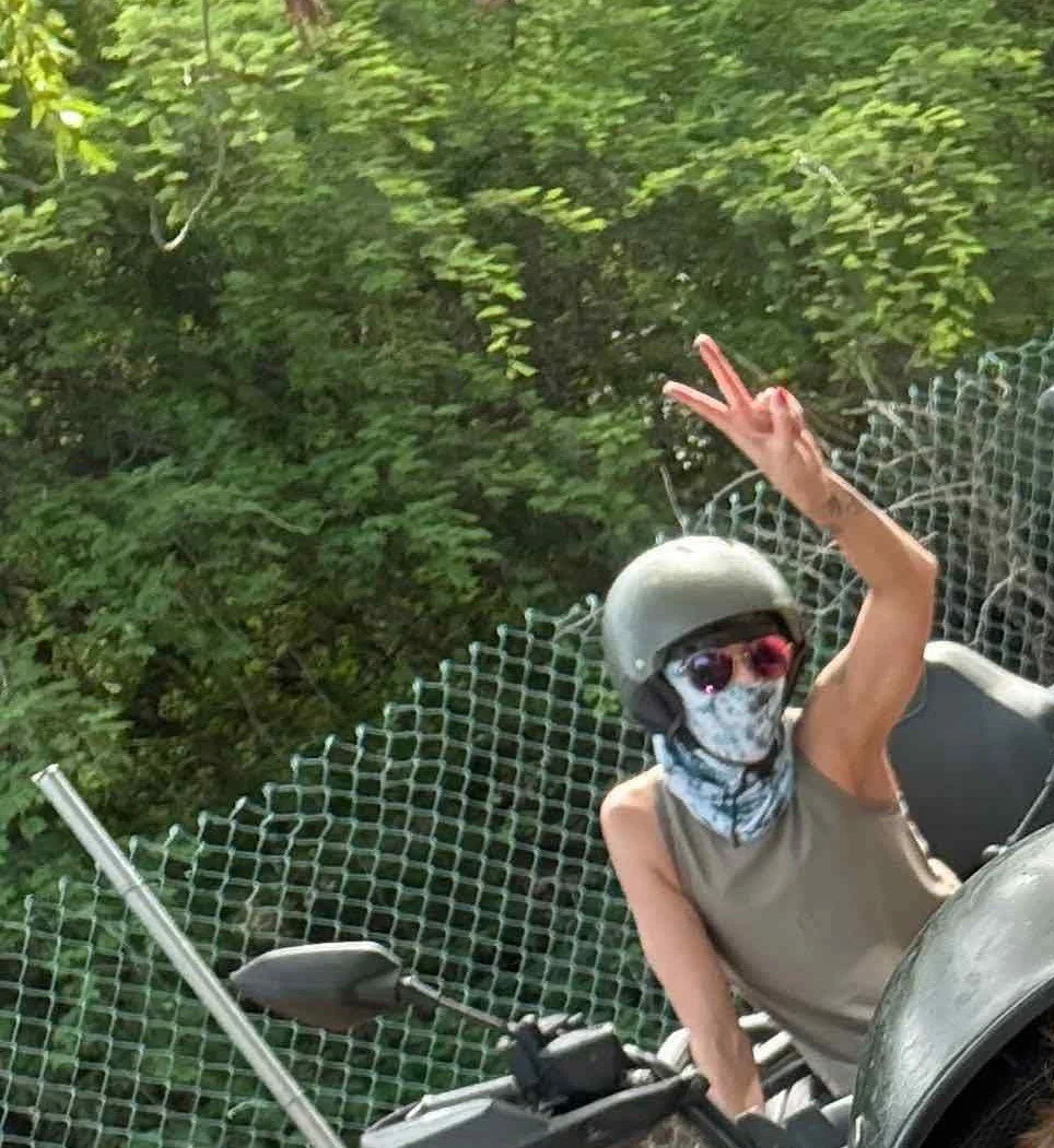 Person wearing a helmet, sunglasses, and face mask riding a motorcycle or scooter, making a peace sign with her hand, with green trees and a chain link fence in the background.