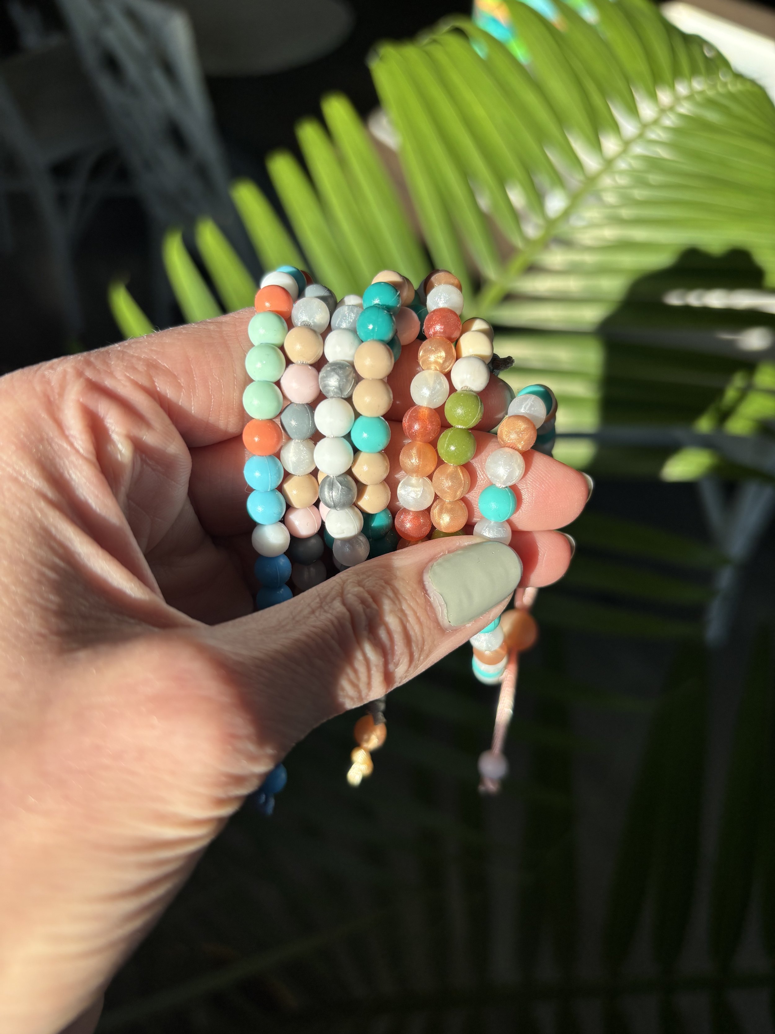 Close-up of a hand holding several colorful beaded bracelets against a backdrop of bright green palm leaves.