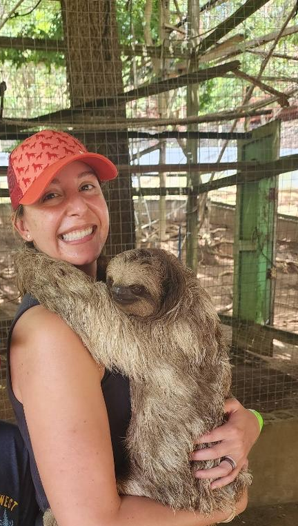 A woman smiling and wearing a red cap with a sloth hanging onto her in an animal enclosure.