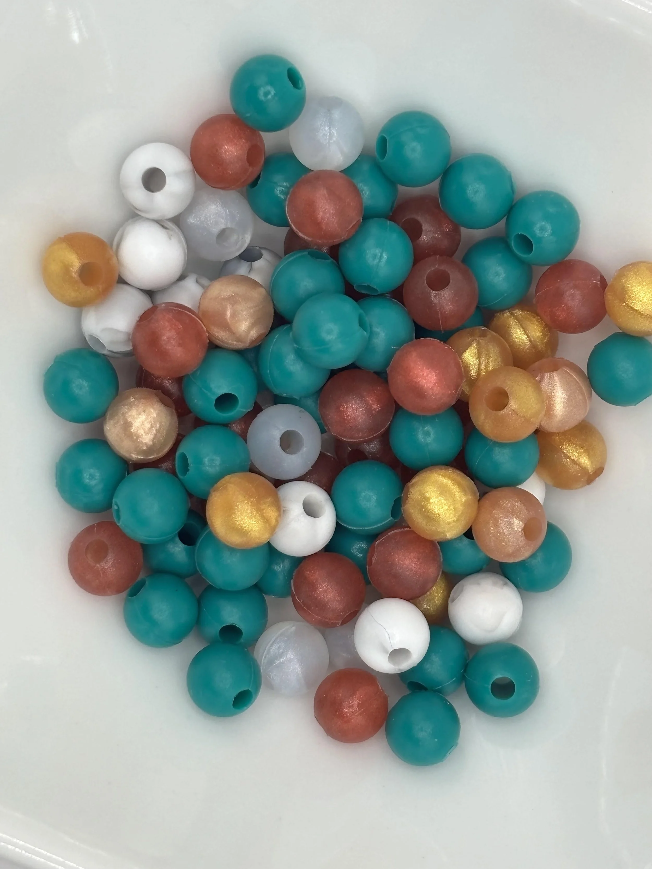 Colorful round beads in teal, white, gold, and bronze in a white bowl.