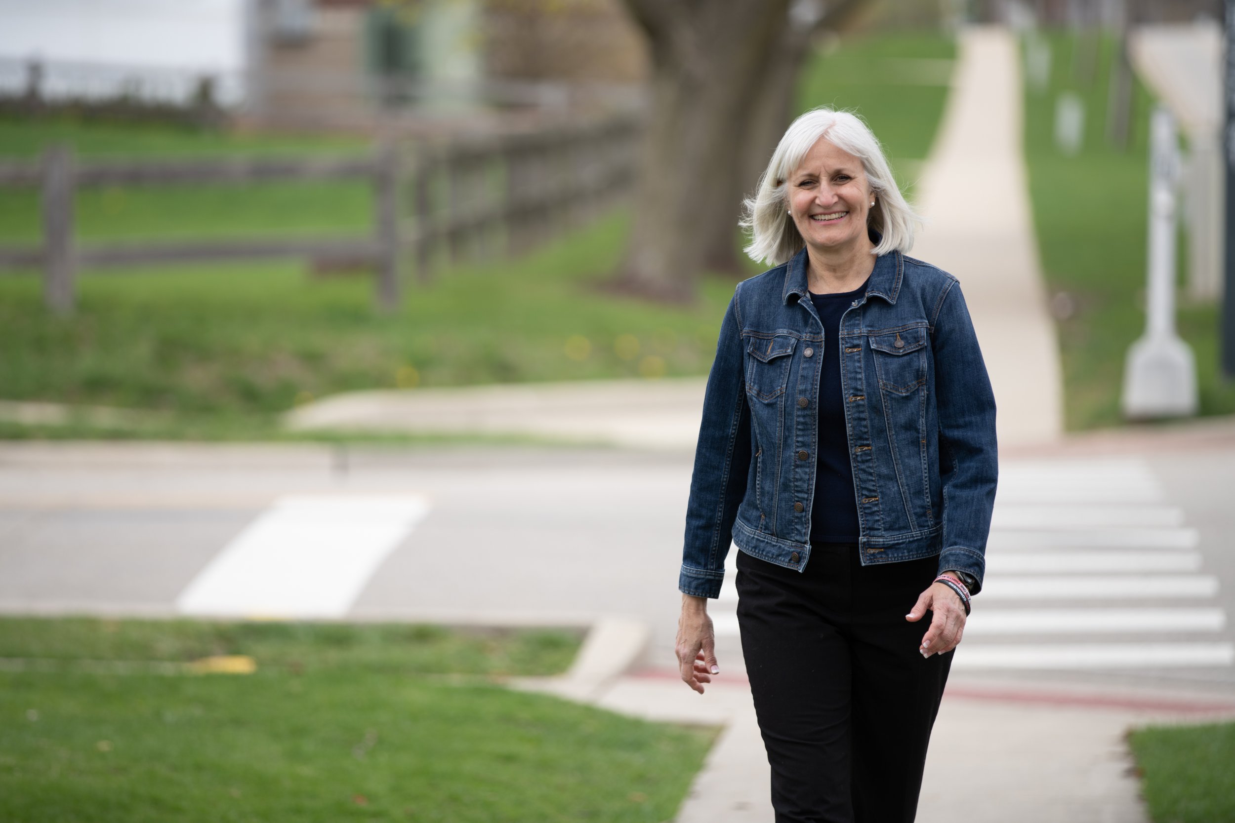 Mary Edly-Allen is smiling and walking on a suburban sidewalk wearing a denim jacket and black pants.