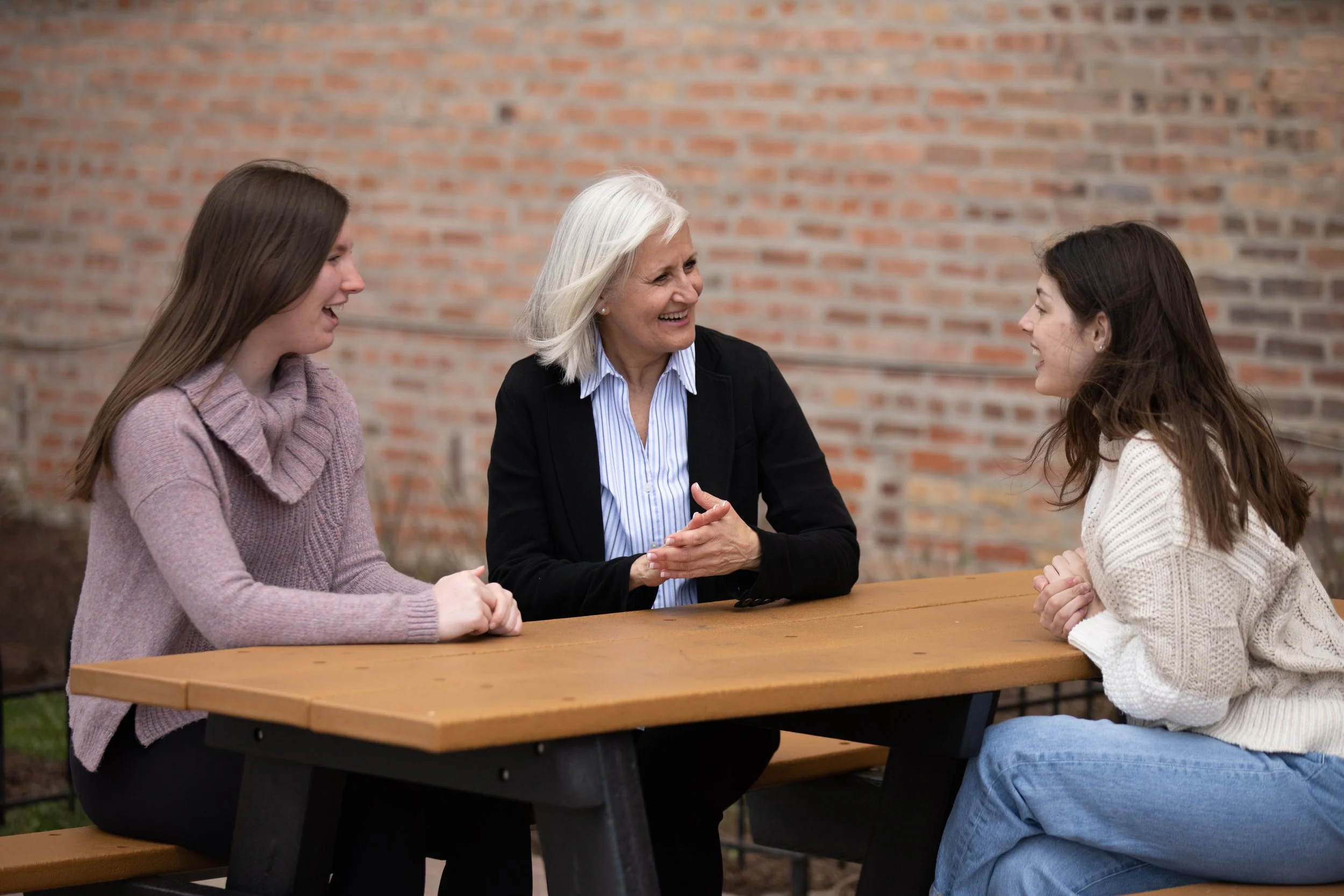 Mary Edly-Allen is with two young women at a picnic table outdoors on a sunny day, having a conversation and smiling.