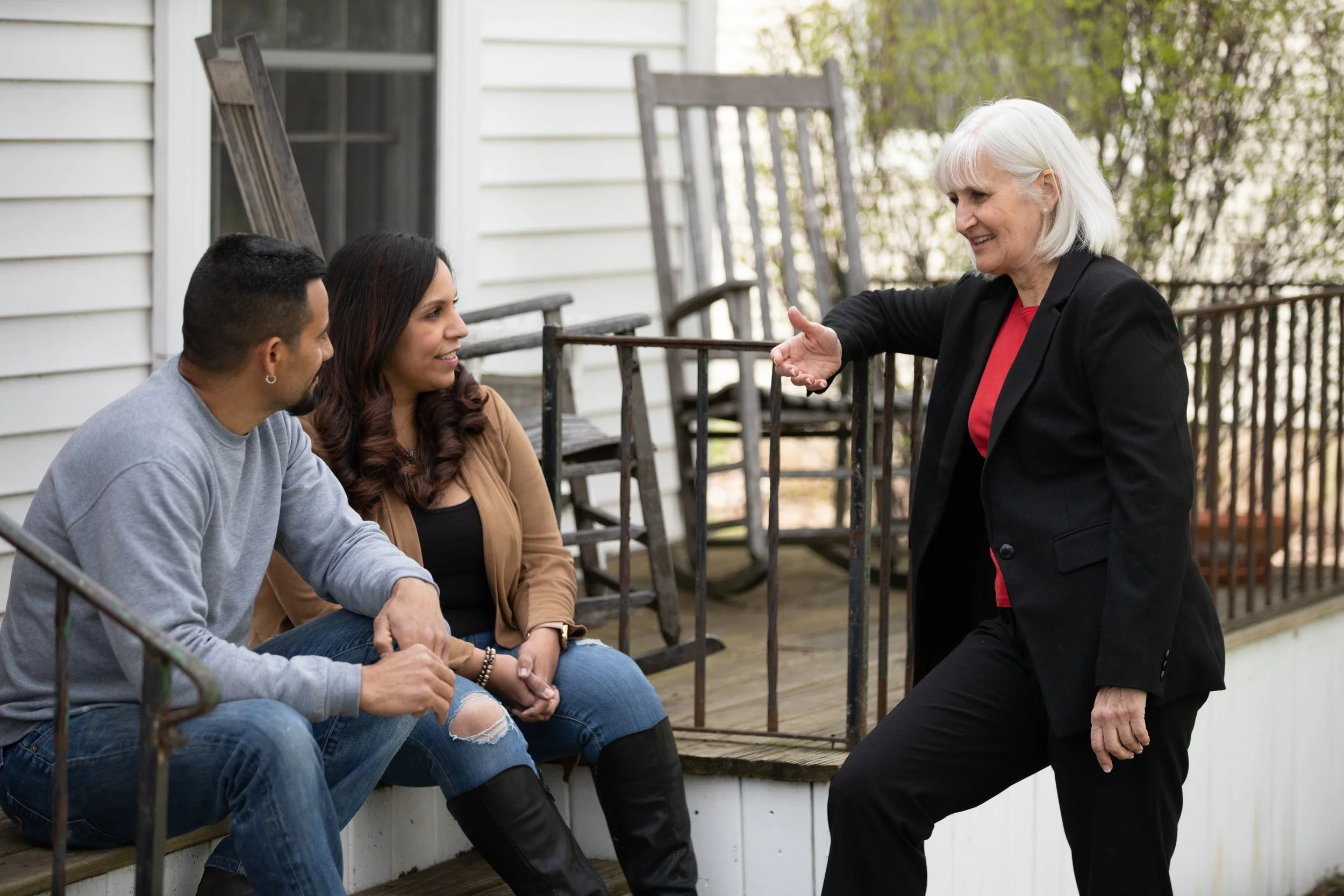 Mary Edly-Allen talking animatedly to a young couple sitting on the porch of a house, all smiling and engaged in conversation.