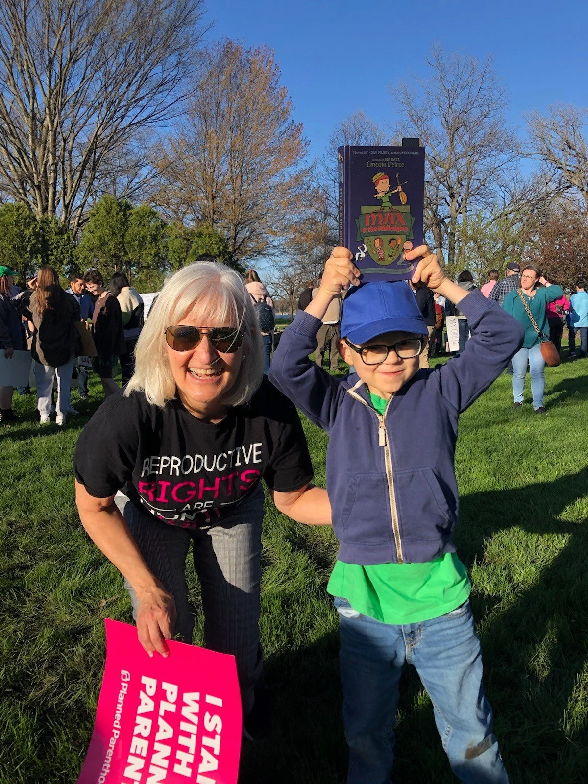 Mary Edly-Allen is wearing sunglasses smiling next to a young boy wearing a blue cap and glasses at an outdoor event. The boy is holding a book above his head. There are many people and leafless trees in the background during a sunny day.