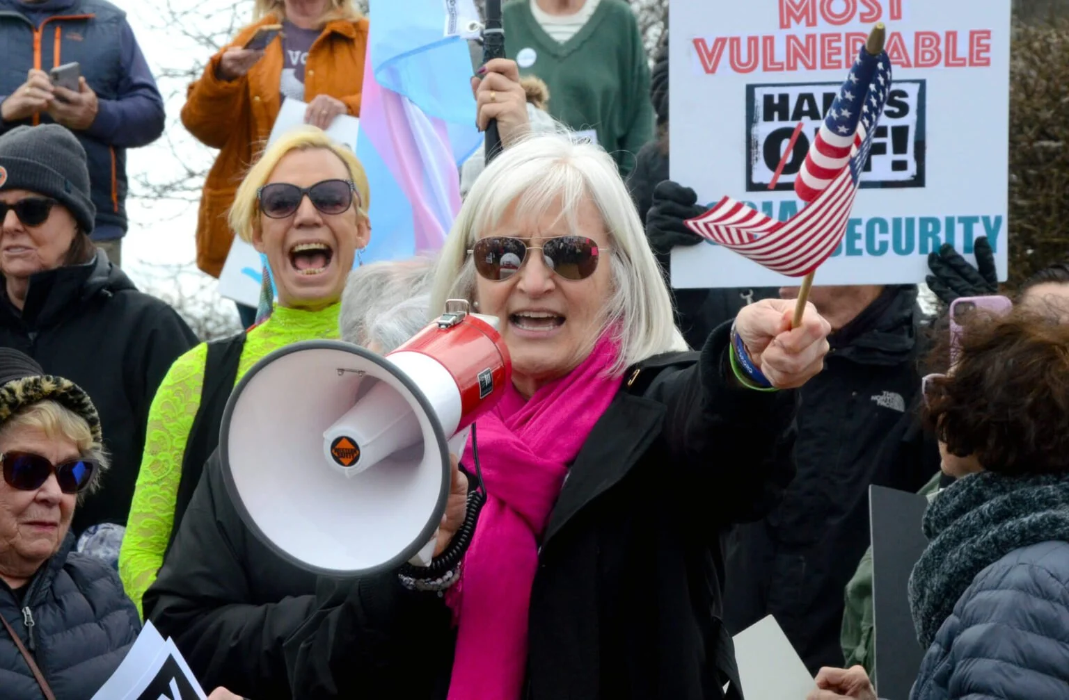 Mary Edly-Allen is wearing sunglasses and a pink scarf holding a small American flag and a megaphone, speaking passionately at a crowded rally.