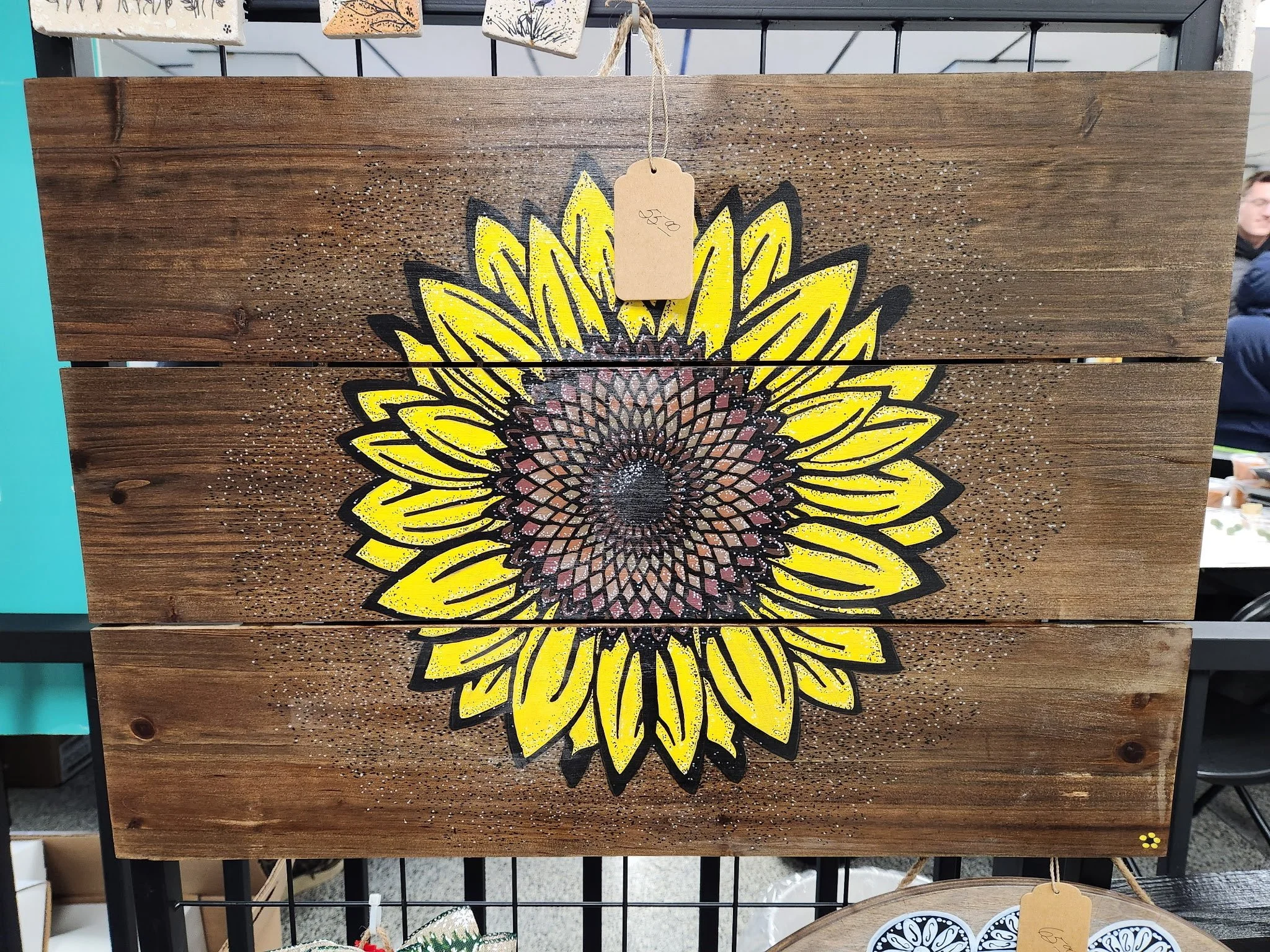Hand-painted sunflower on wooden board with yellow petals and detailed center, hanging on display rack at a store.