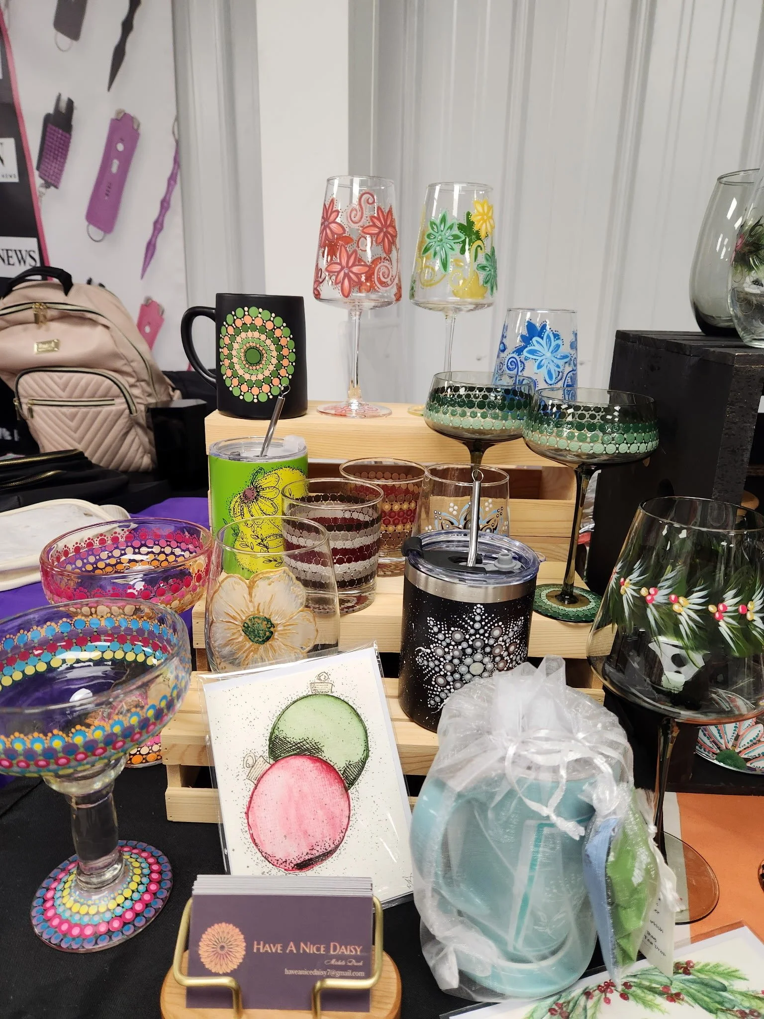 Colorful handcrafted glassware and decorative items on display at a market or craft fair, including painted glasses, a black mug with painted dots, and small artworks.