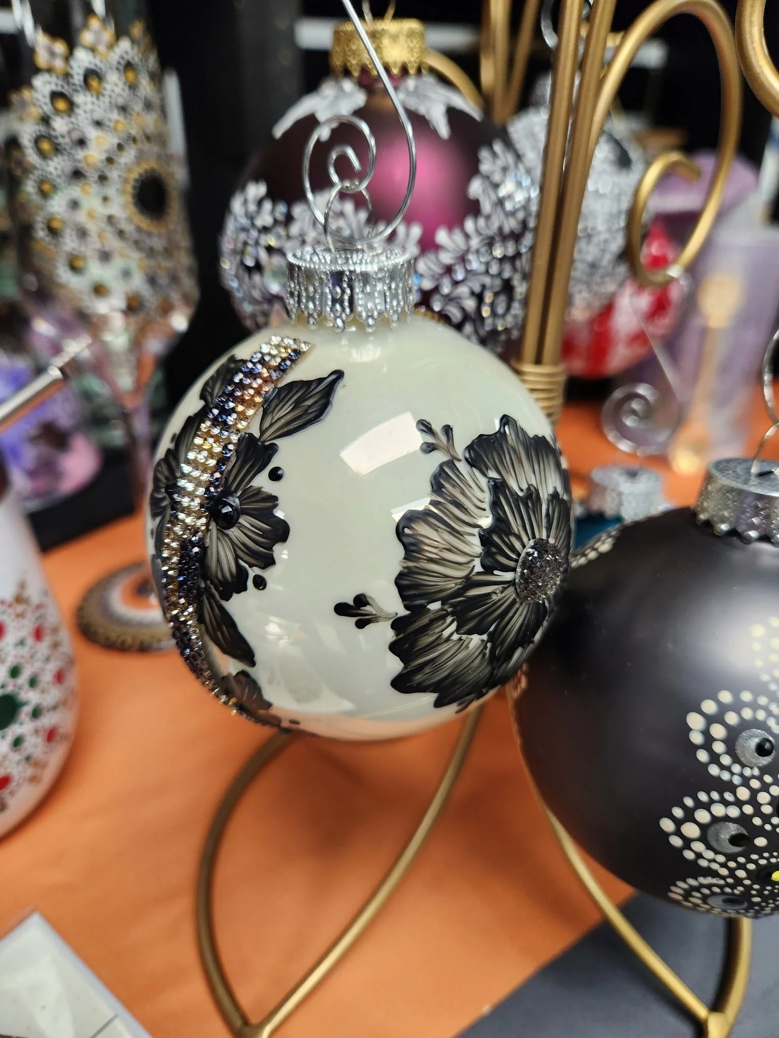Decorative Christmas ornaments on display, including a white ornament with black floral designs and rhinestones, surrounded by other shiny, colorful ornaments.