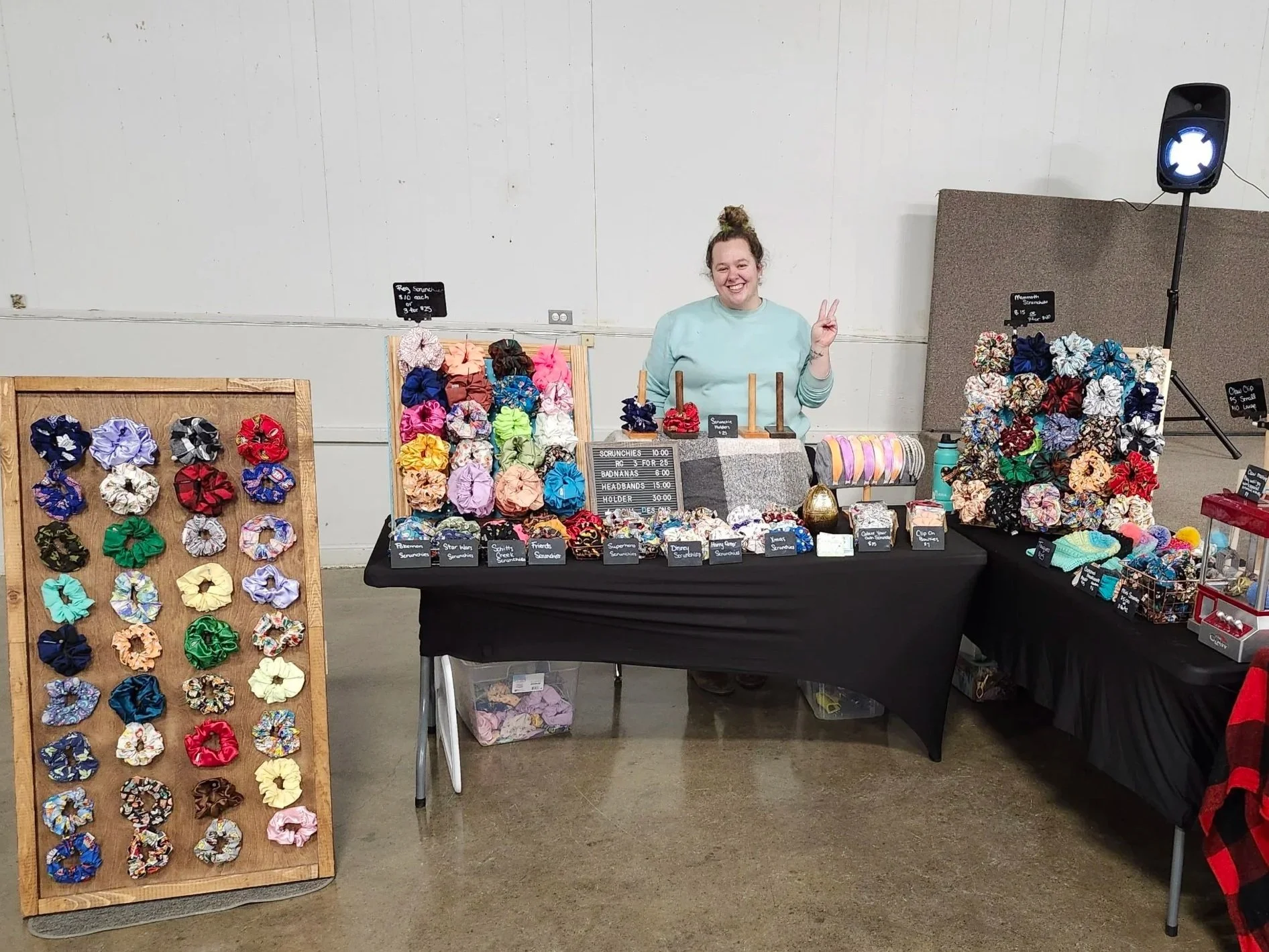 A woman smiling and making a peace sign behind a table selling colorful scrunchies, headbands, and hair accessories at an indoor market or craft fair.
