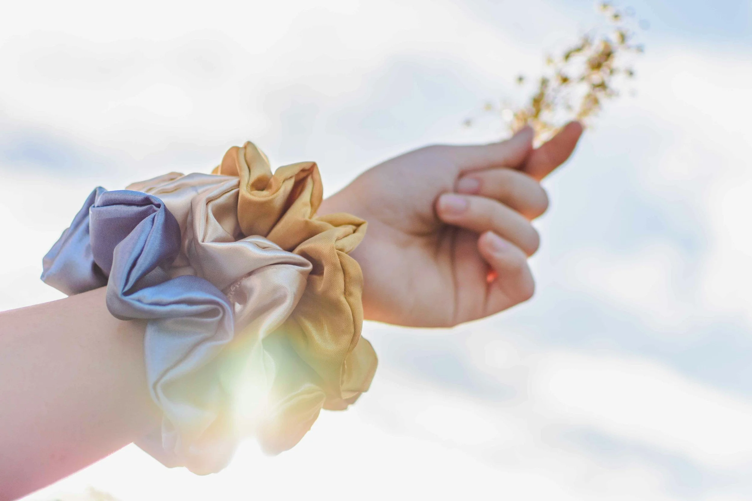 A person's hand with a mix of beige, yellow, and pastel blue scrunchies on the wrist, reaching towards a bright, partly cloudy sky, holding a small bunch of flowers.