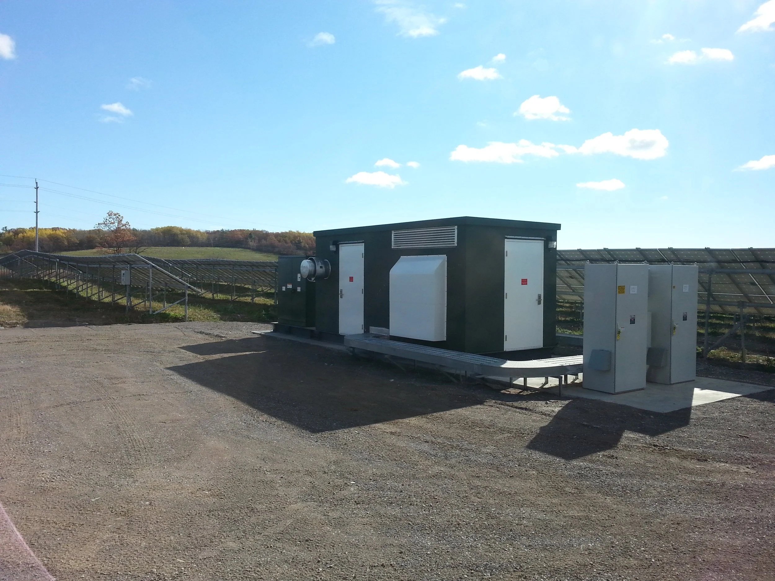 A solar panel installation with electrical equipment in a rural area under a blue sky with some clouds.