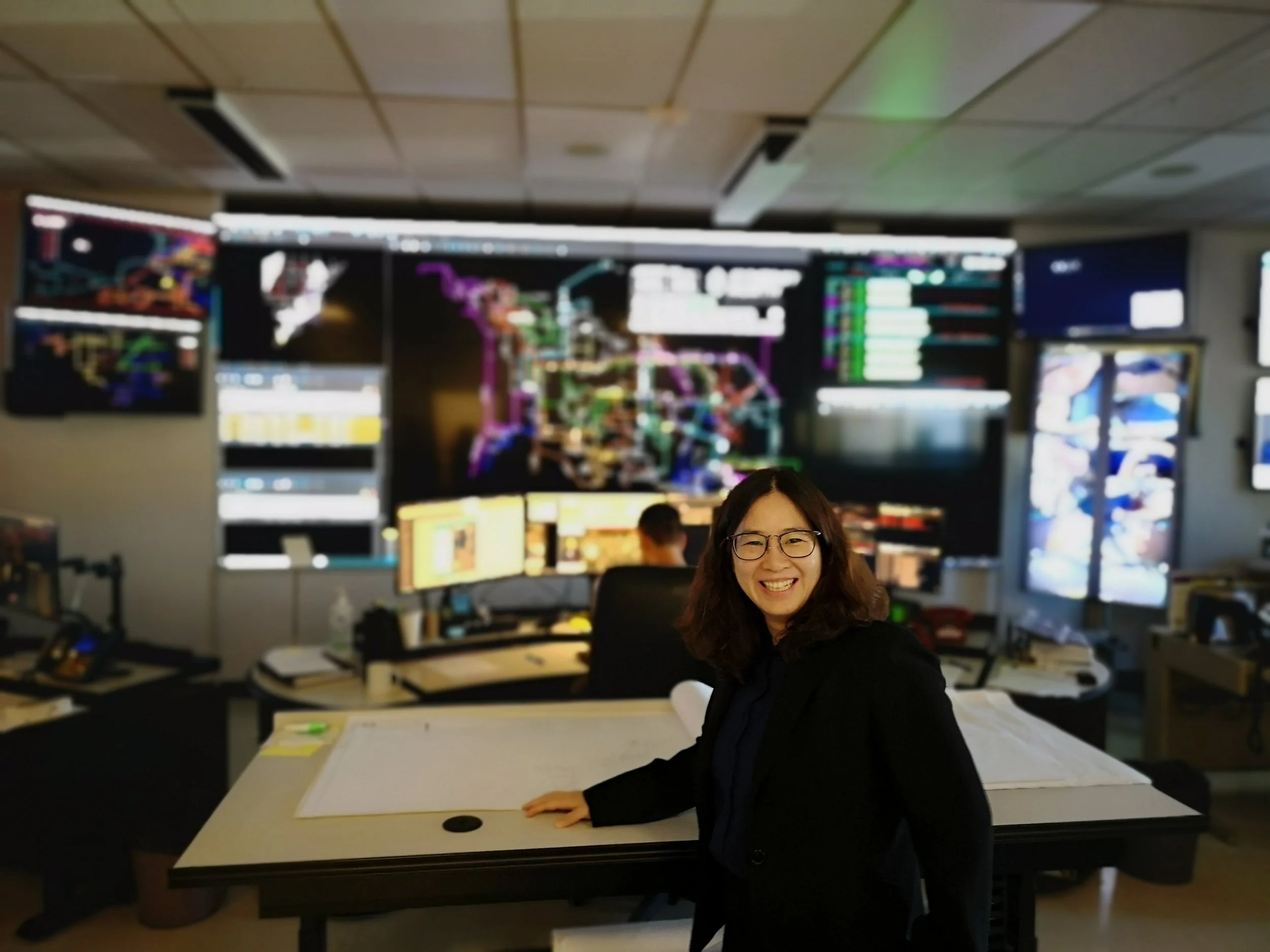 A smiling woman with glasses in an office or control room with multiple computer screens displaying colorful graphs and data.