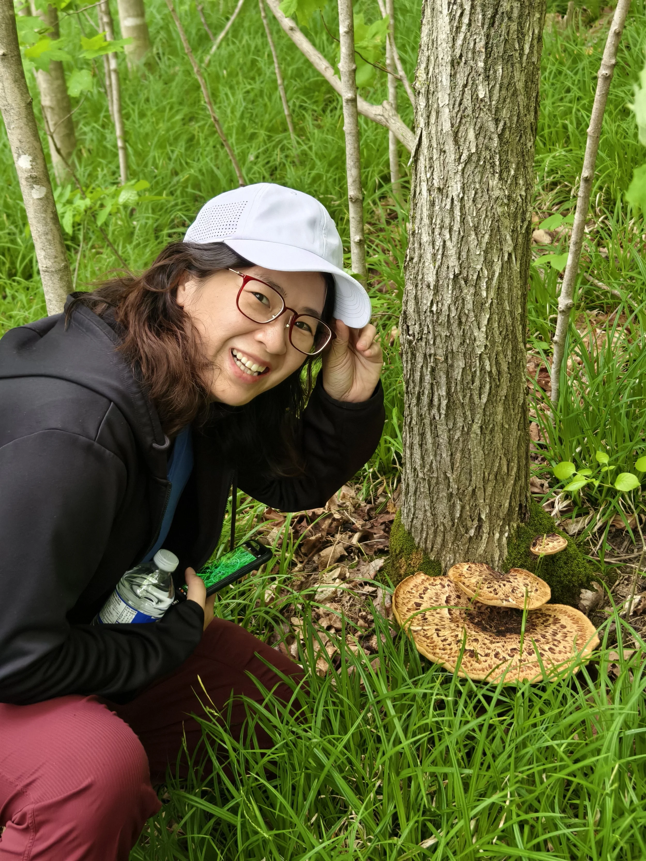 A woman smiling, wearing glasses, a white cap, black jacket, and reddish-brown pants, crouching next to a tree with large mushrooms at its base, holding a water bottle and a smartphone in a lush green forest.
