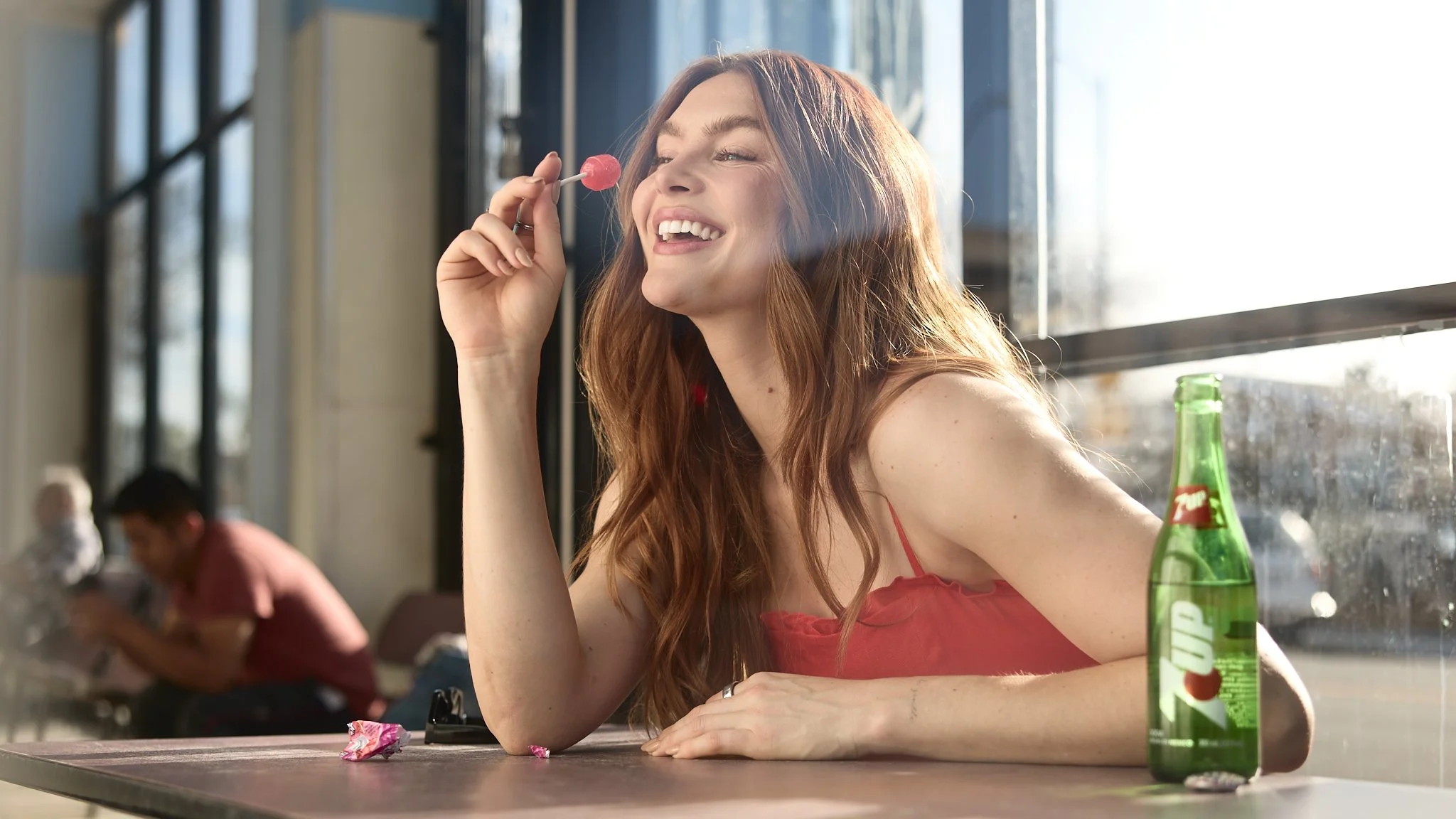 A smiling red-haired woman sitting at a table, holding a pink lollipop, with a green glass bottle of 7UP soda on the table, in a bright indoor setting with large windows.