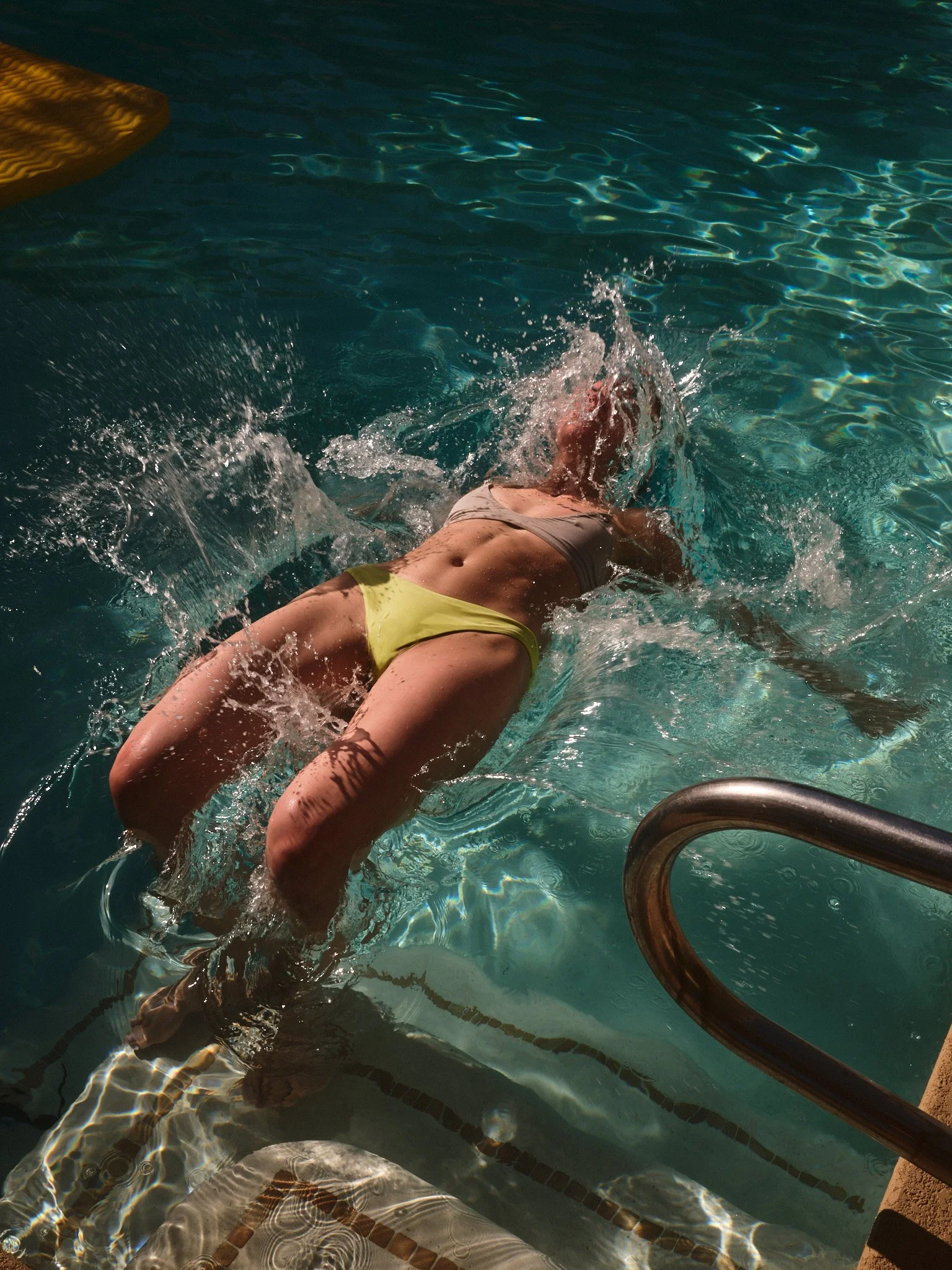 Person wearing a yellow swimsuit and a white top entering a swimming pool using a metal ladder.