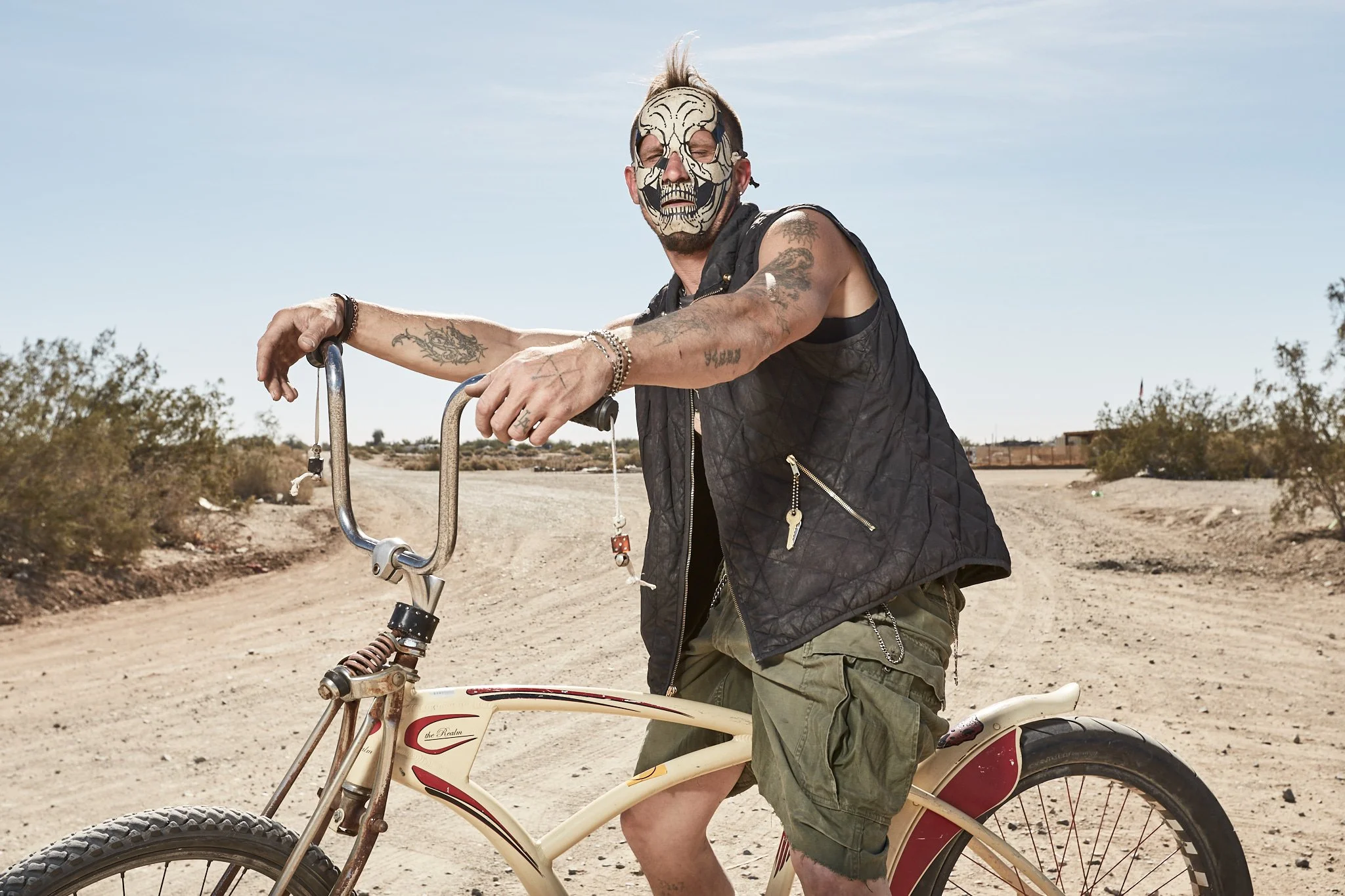 A man with tattoos, wearing a black vest, green shorts, and a skull mask, leans on a vintage bicycle in a desert landscape.