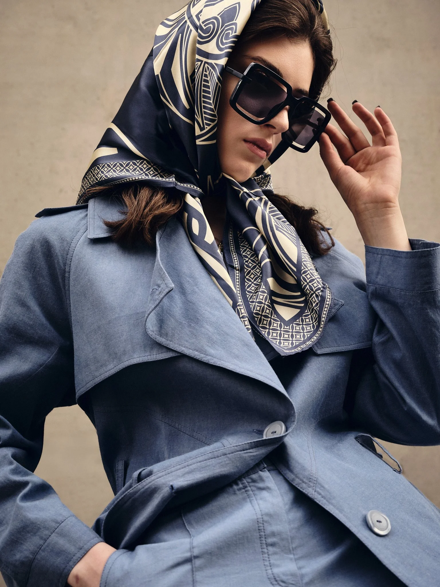 Fashionably dressed woman with large sunglasses, a patterned scarf on her head, and a light gray trench coat, posing against a plain background.