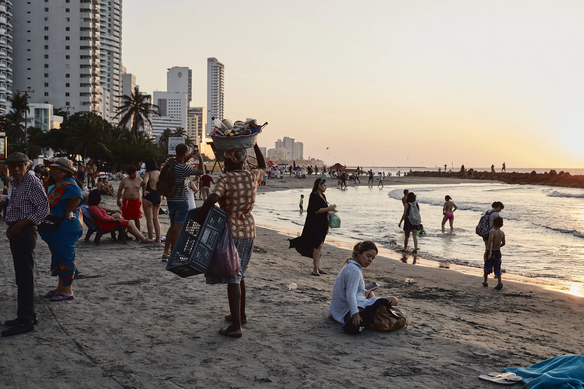 Beach scene with people walking, sitting, and swimming at sunset, with city buildings in the background.